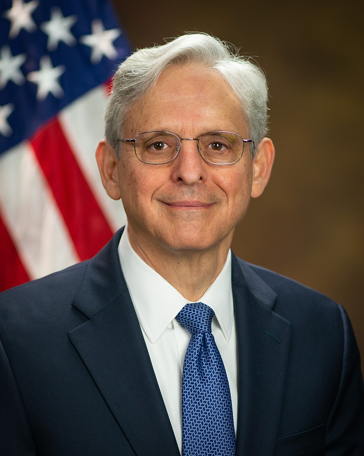 A man with short gray hair, wearing a navy suit, white shirt, and blue tie, with a blurred American flag in the background.