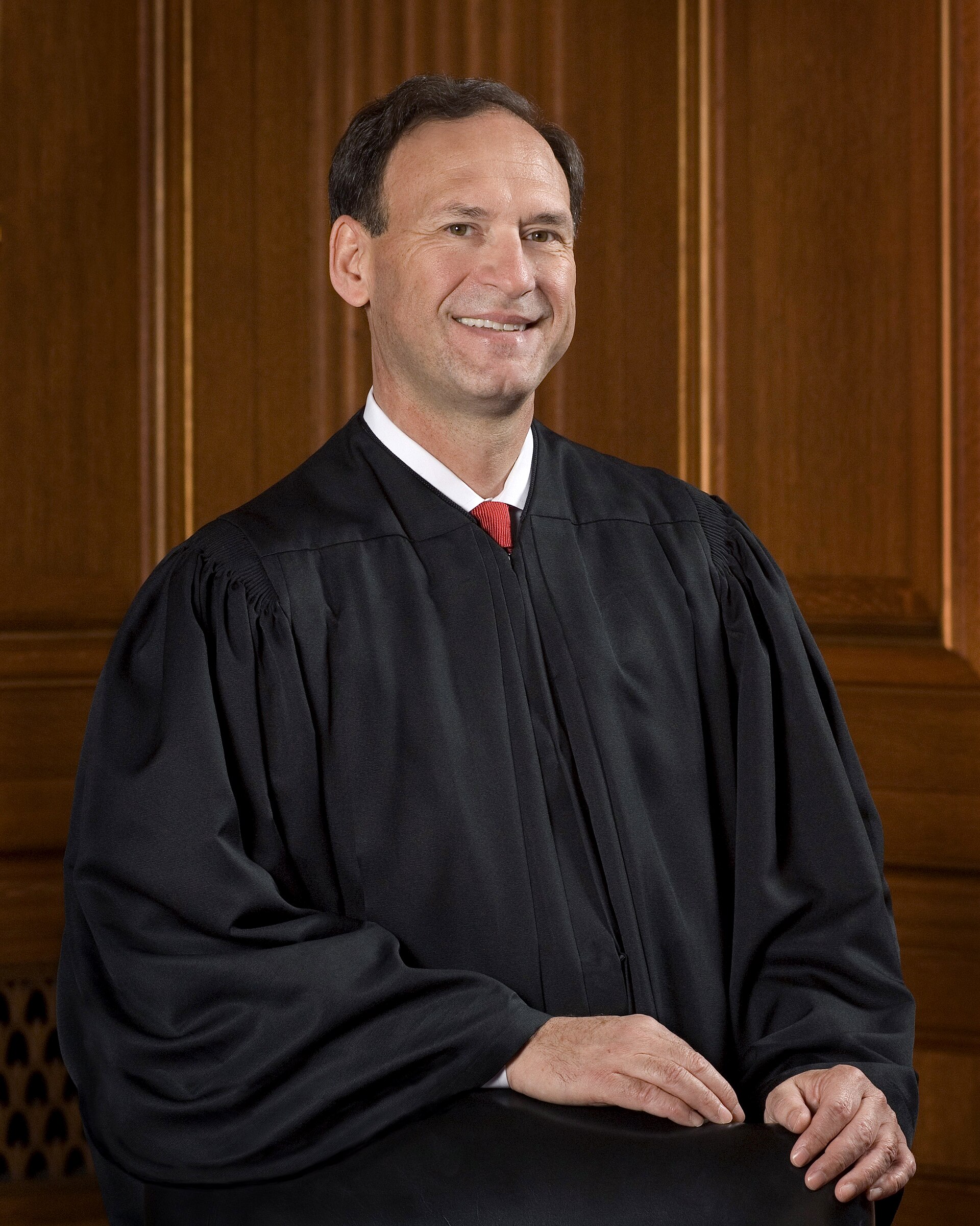 A middle-aged man with short dark hair, wearing a black judicial robe, a white collared shirt, and a red tie, sits in front of a brown wooden wall, smiling and looking to the right.
