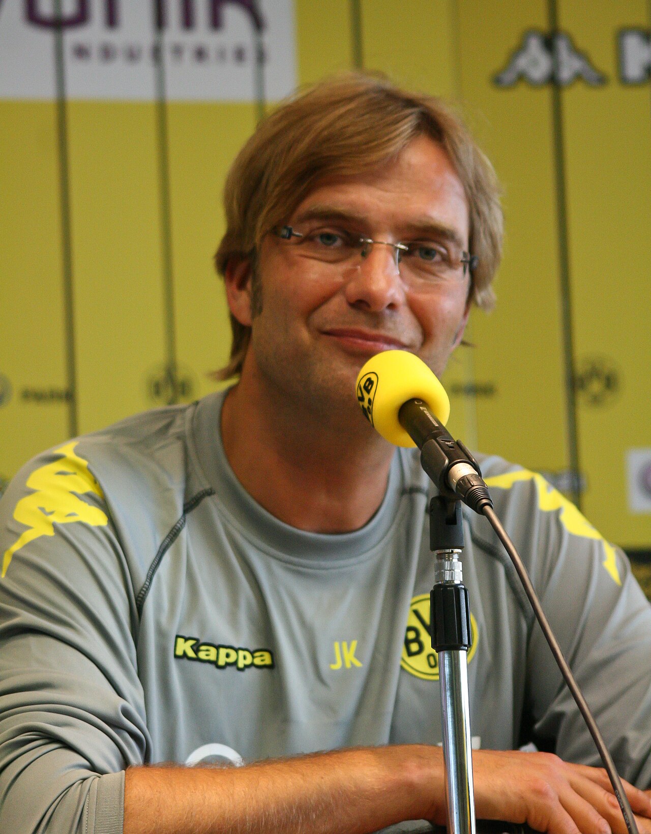 A man with blond hair and glasses wearing a gray shirt with yellow accents and a microphone in front of him.