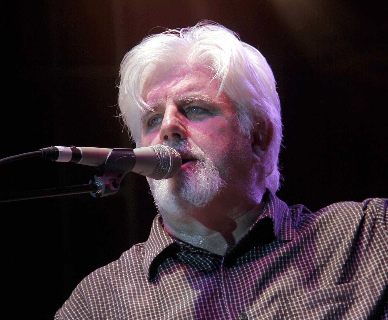 A man with white hair and a beard, wearing a plaid shirt, singing into a microphone.