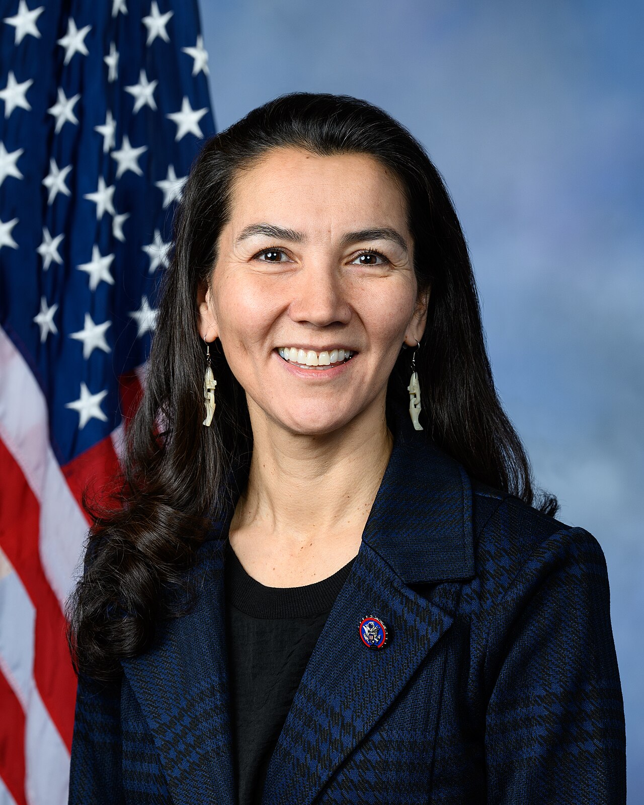 Mary Peltola, a woman with long dark hair, smiles and wears a dark blue plaid jacket over a black shirt. She has on earrings and a pin on her lapel. Behind her is an American flag.