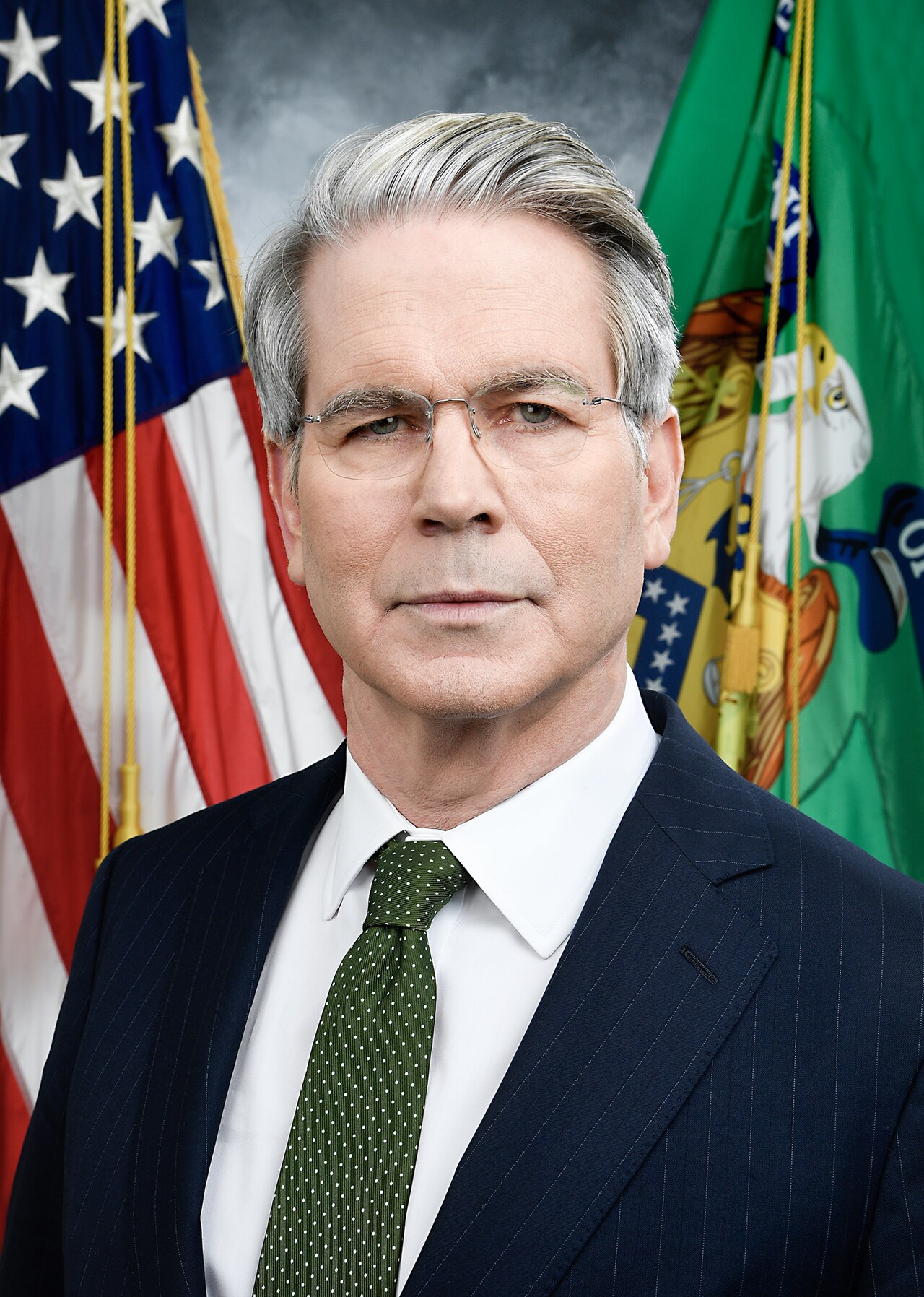 A man in a suit and tie stands in front of the American flag and another green flag.