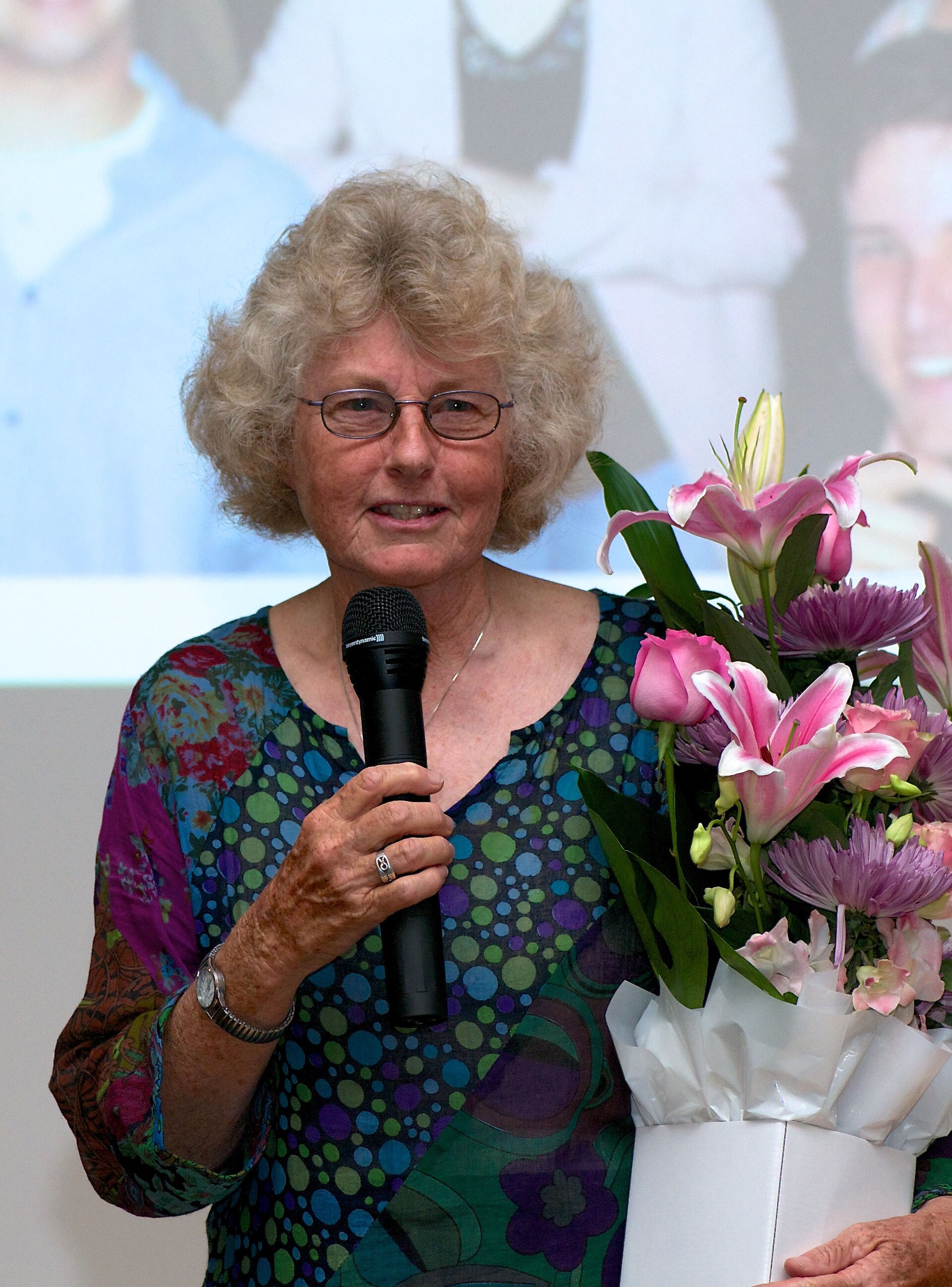 A woman with short, curly blonde hair and glasses holding a microphone and a bouquet of flowers.