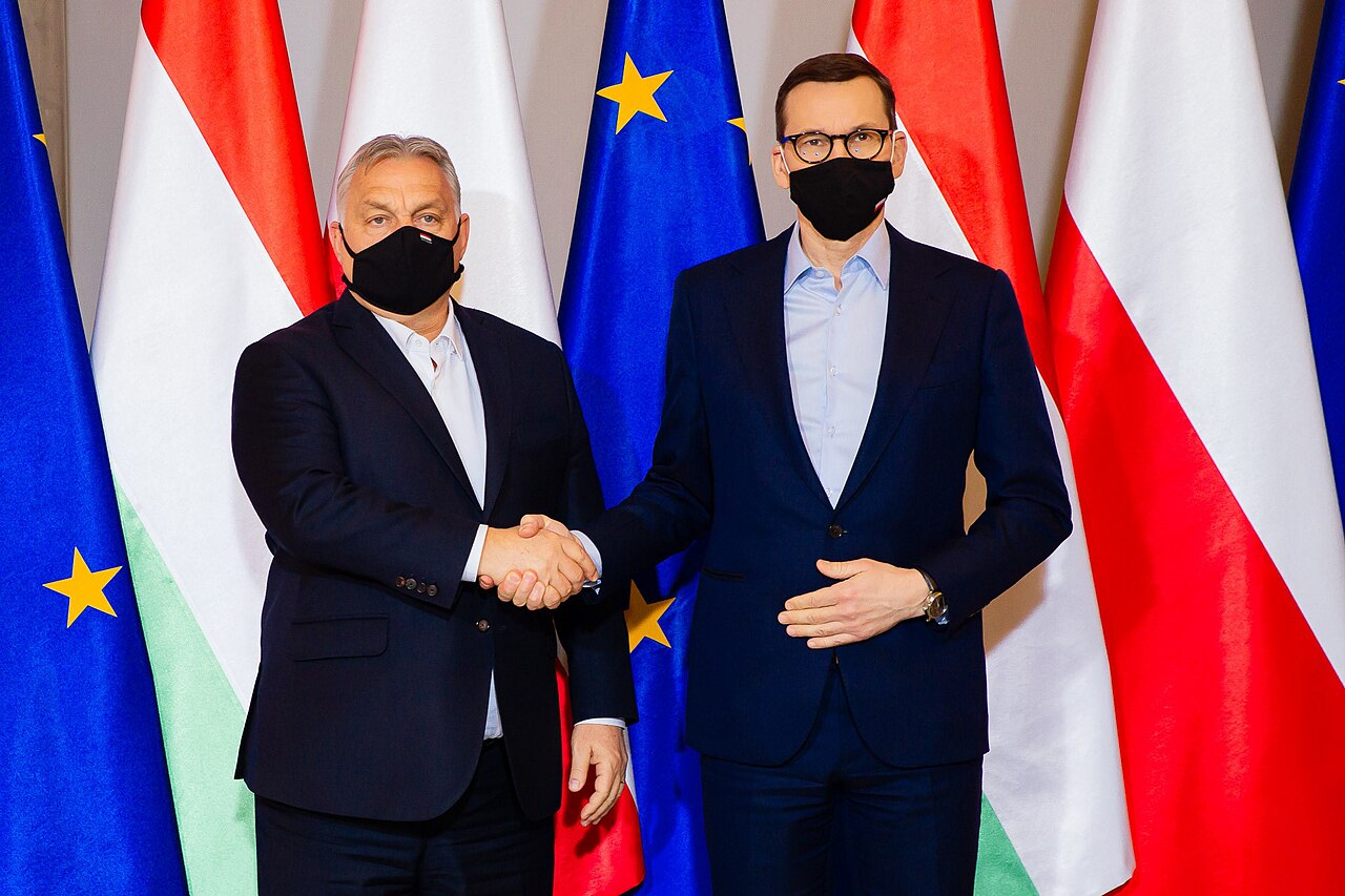 Two men in suits and face masks shake hands in front of national flags.
