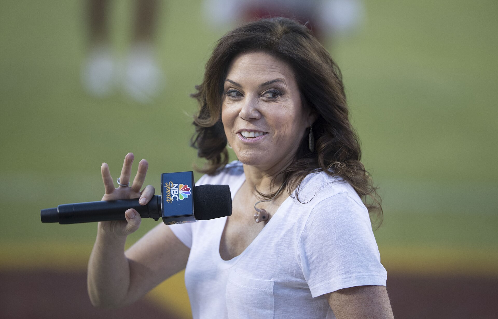 Michele Tafoya, a woman with long brown hair, wearing a white t-shirt, holds an NBC Sports microphone in her right hand.