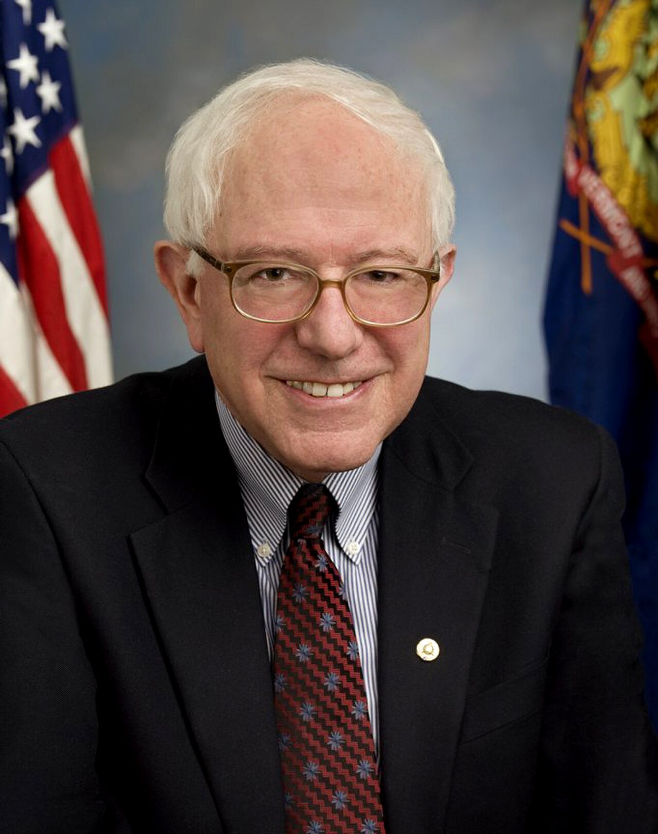 Bernie Sanders smiling and wearing a suit, with two flags behind him.