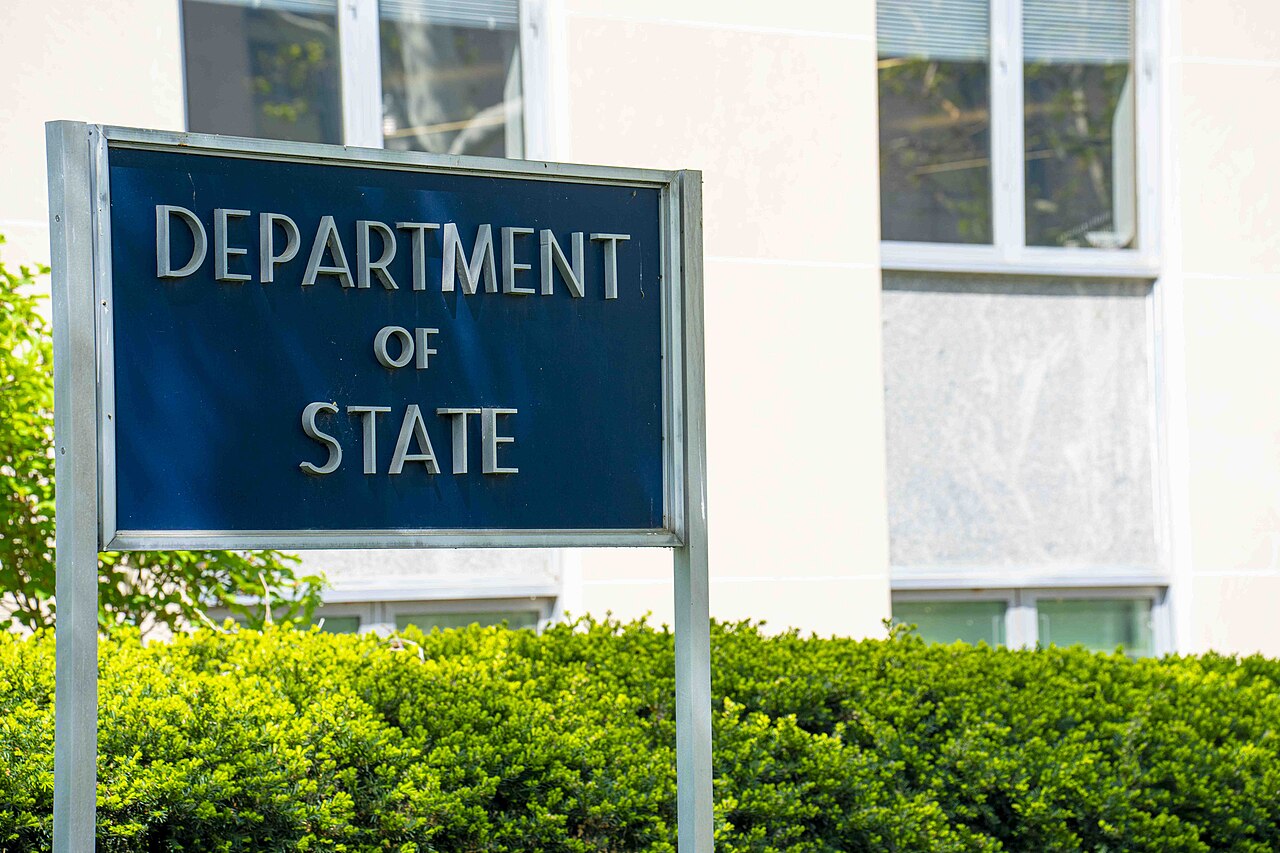 A blue sign with silver lettering that reads DEPARTMENT OF STATE in front of a building and green bushes.