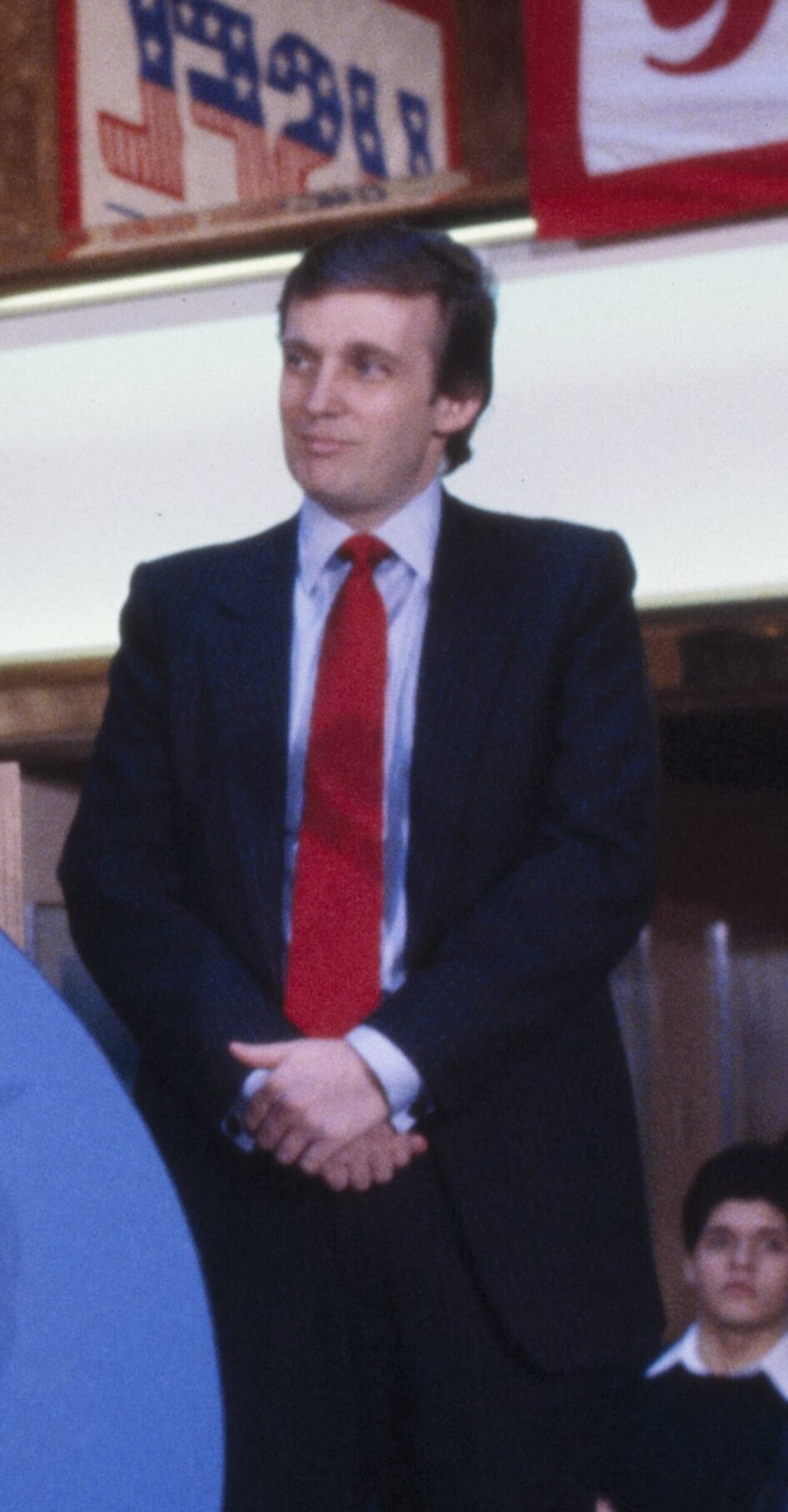 Donald Trump in a dark suit and red tie stands with his hands clasped in front of him.