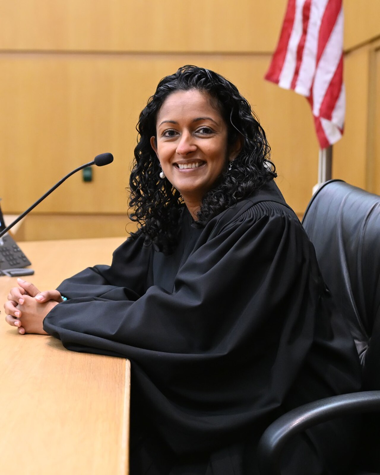 A female judge in a black robe sitting at a desk with her hands clasped together in front of her.