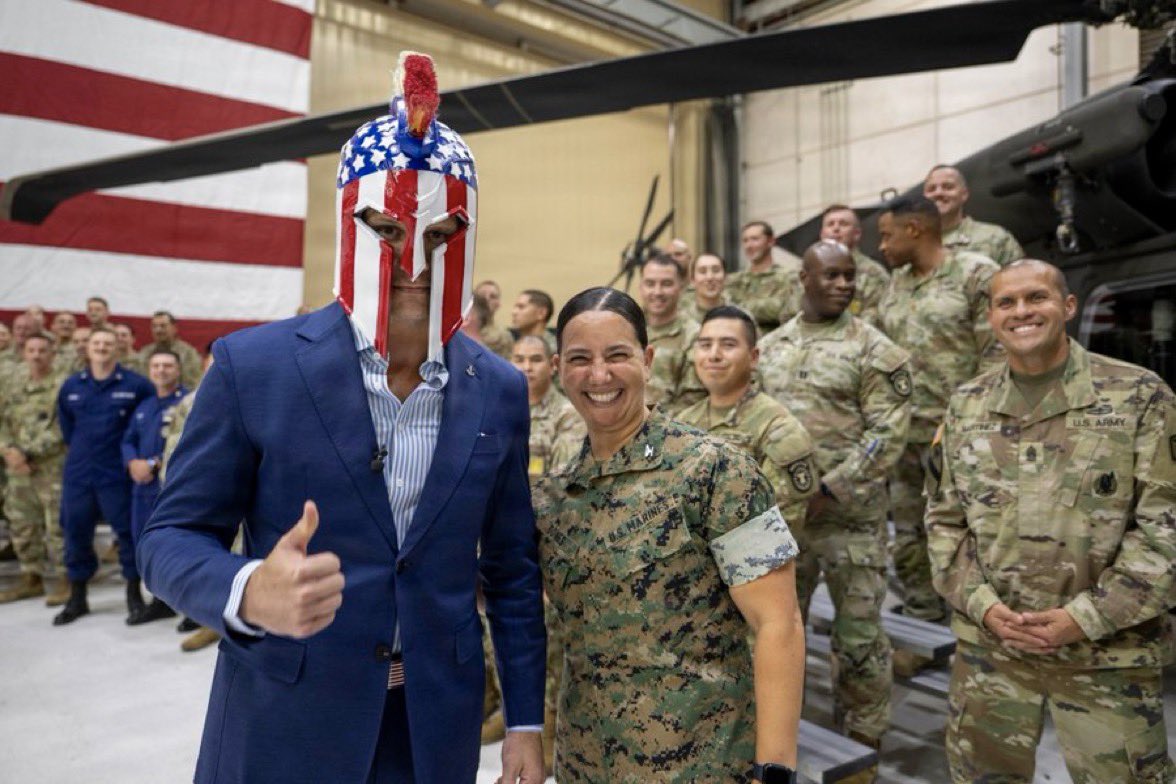 Pete Hegseth in a blue suit giving a thumbs-up beside a Marine in a hangar, with a stars-and-stripes Corinthian helmet and red plume on his head. A flag and a helicopter are behind them.