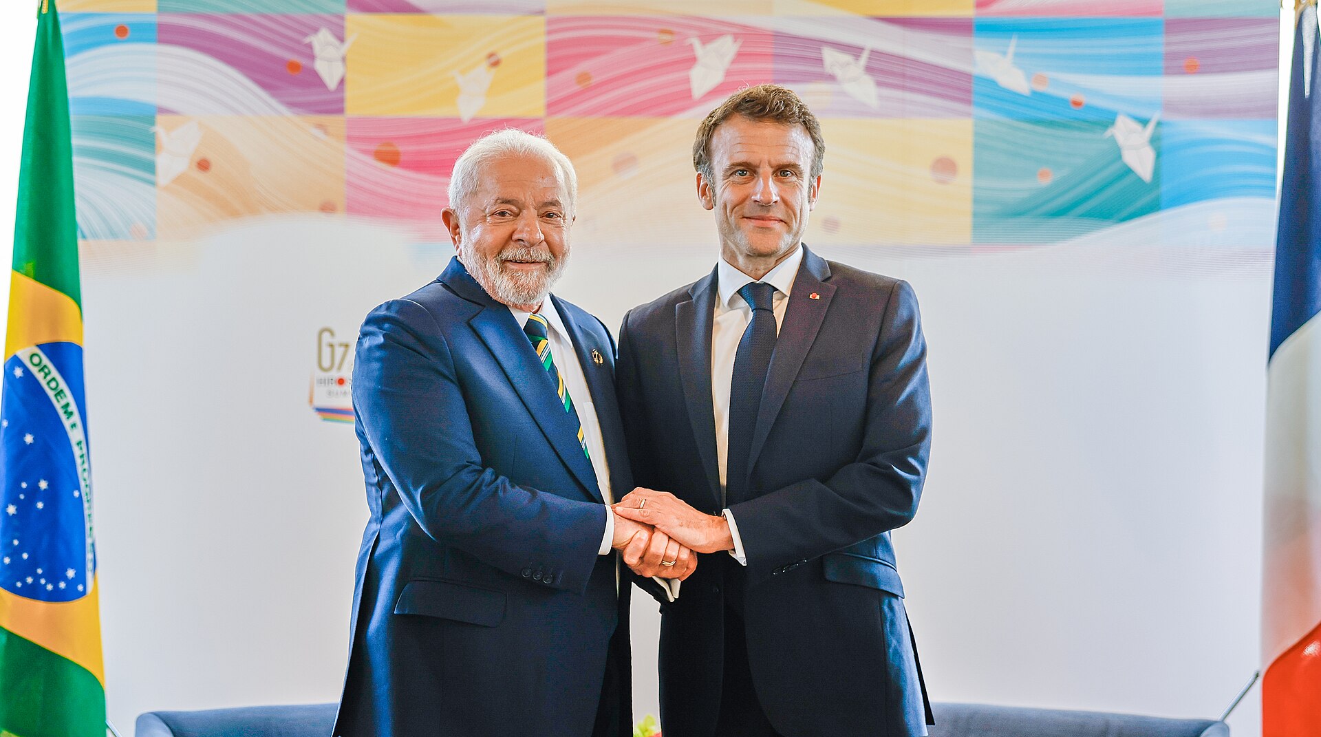 Two men in suits shaking hands in front of flags and a colorful backdrop.