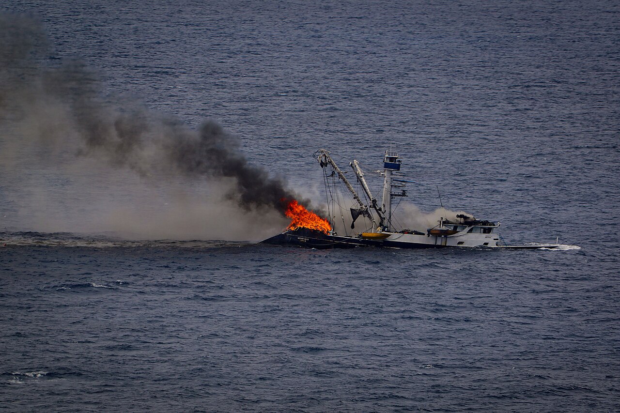 A fishing boat is on fire in the ocean, with thick black smoke billowing from its deck and flames engulfing part of the vessel.