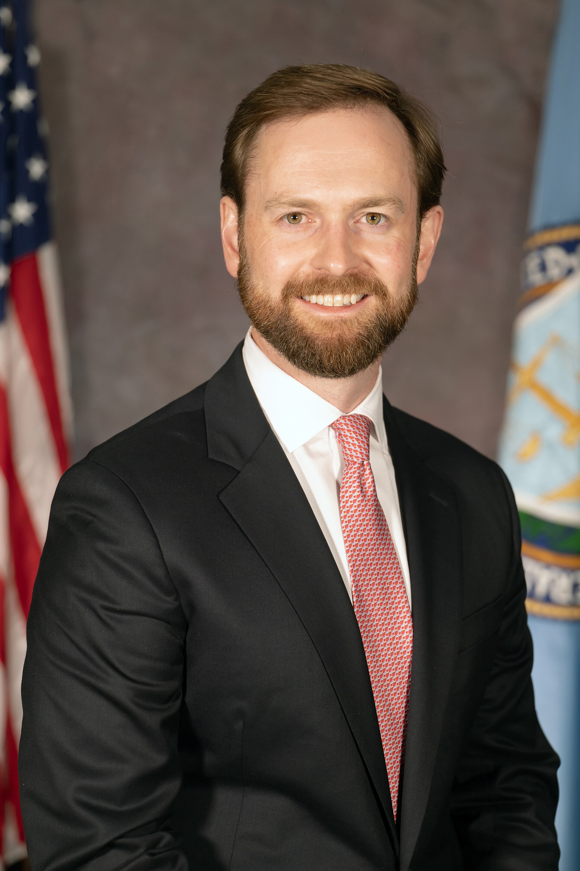 A smiling man in a black suit, white shirt, and red patterned tie, with an American flag and another flag behind him.
