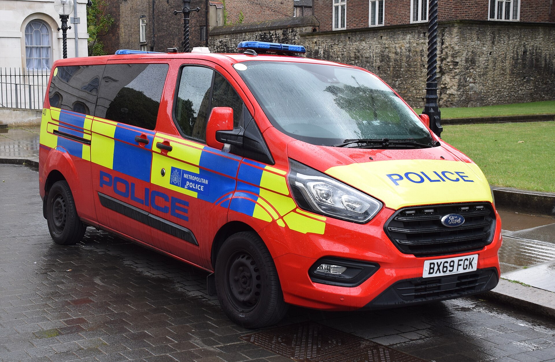 A red and yellow Metropolitan Police van with blue flashing lights is parked on a wet, paved street. The word "POLICE" is written in blue on the side and hood of the van. The license plate reads BX69 FGK.