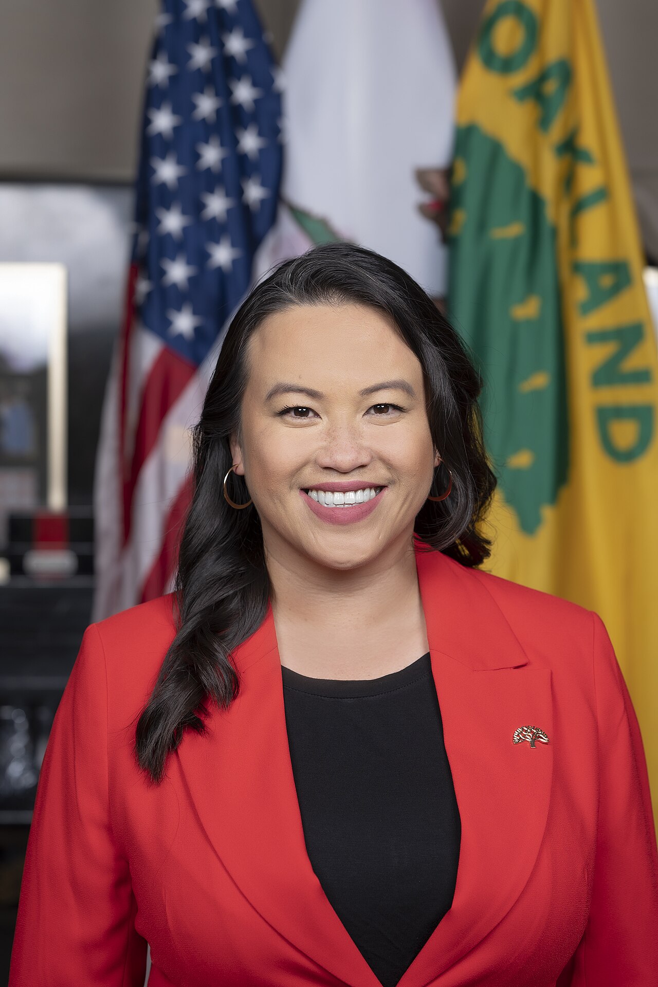 A woman in a red blazer and black top smiles at the camera, with the American flag, a white flag, and a yellow flag with green text and graphics behind her.