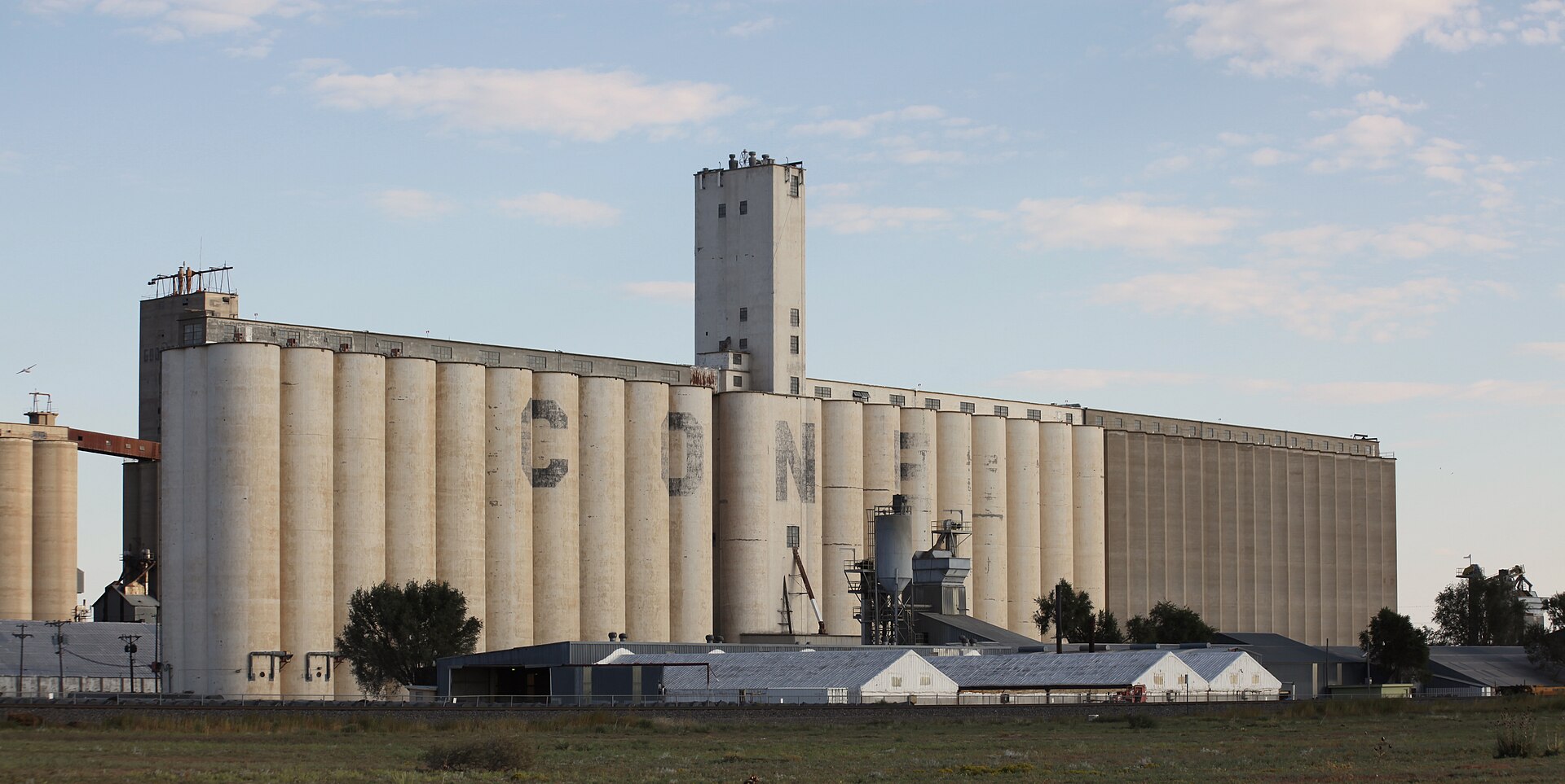 A long row of concrete grain silos with the letters "CON AG" visible on them, under a partly cloudy sky.