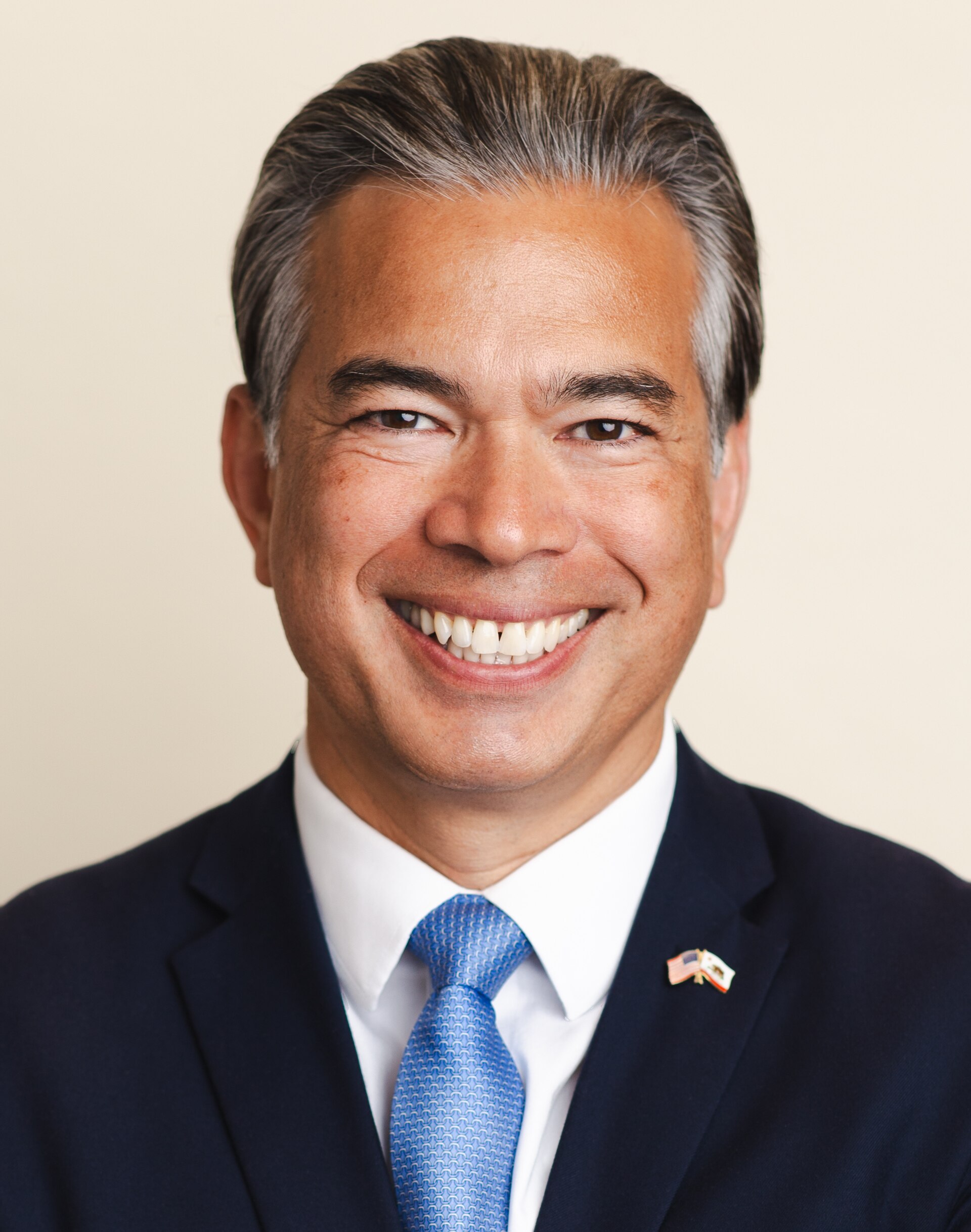 A smiling middle-aged man with short graying hair, wearing a dark suit, white shirt, blue tie, and a small pin on his lapel, against a beige background.