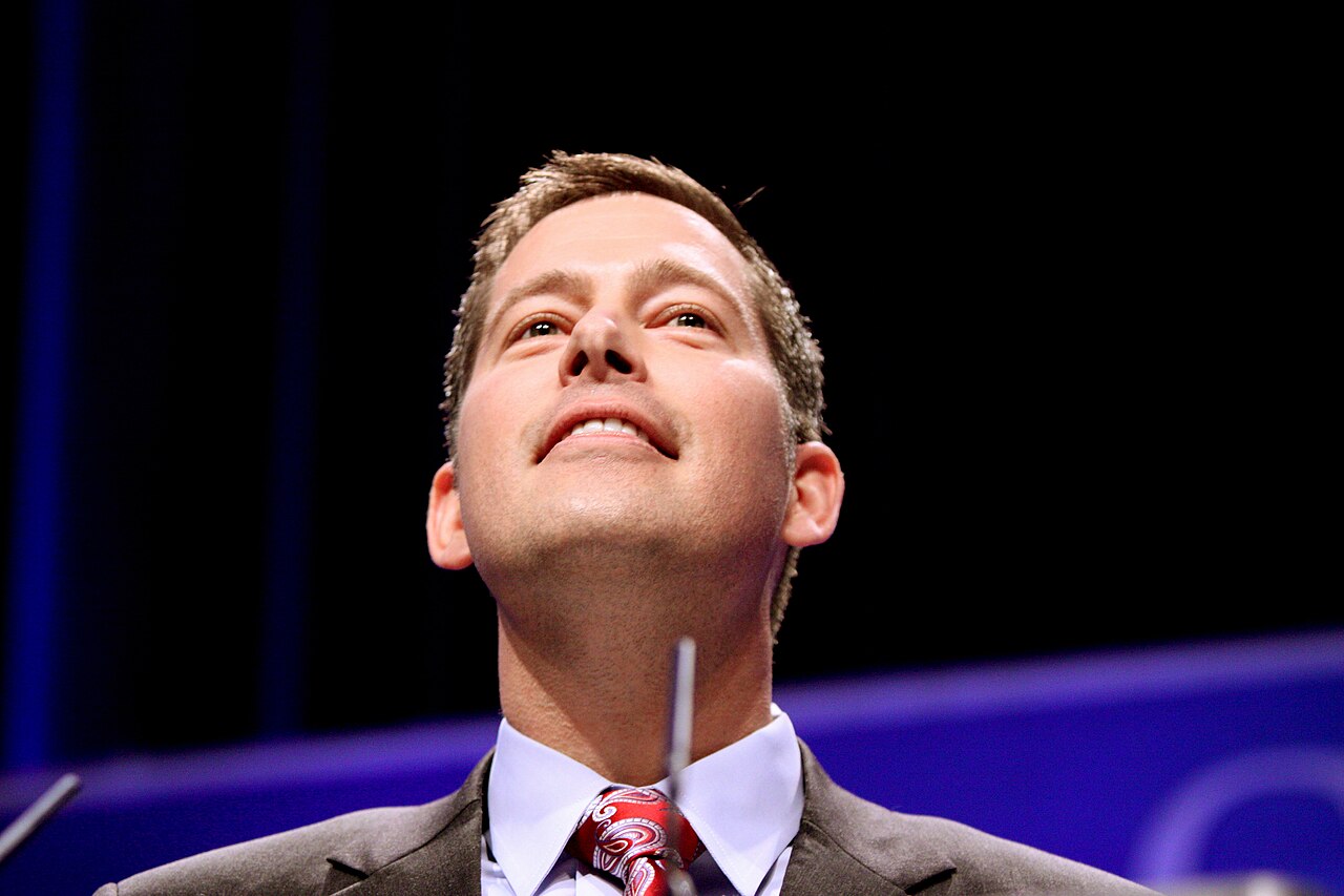 a fair-skinned man in a gray suit and red tie looks up