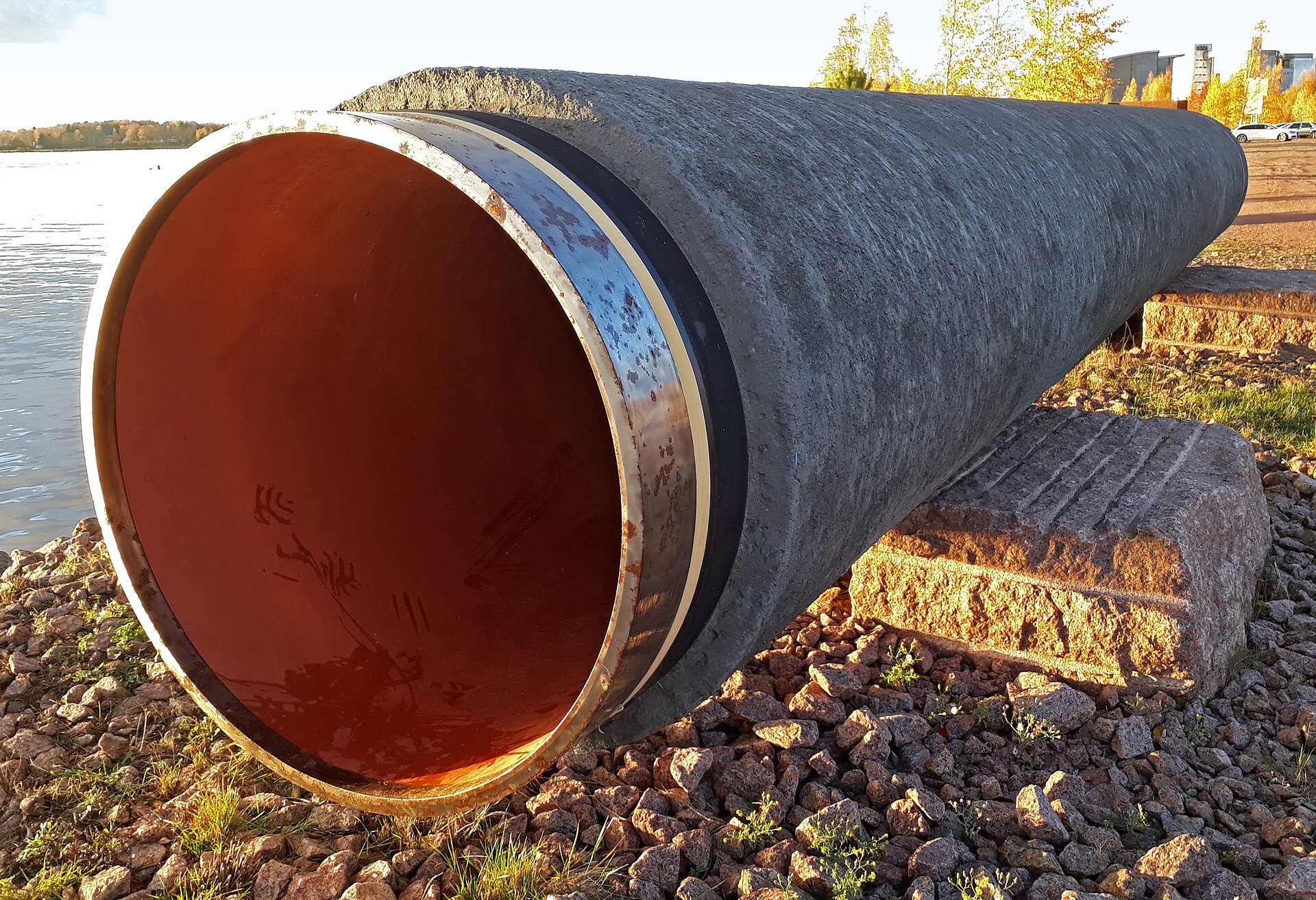 A large, grey, concrete pipe lies on its side on a rocky shore, with a body of water in the background.