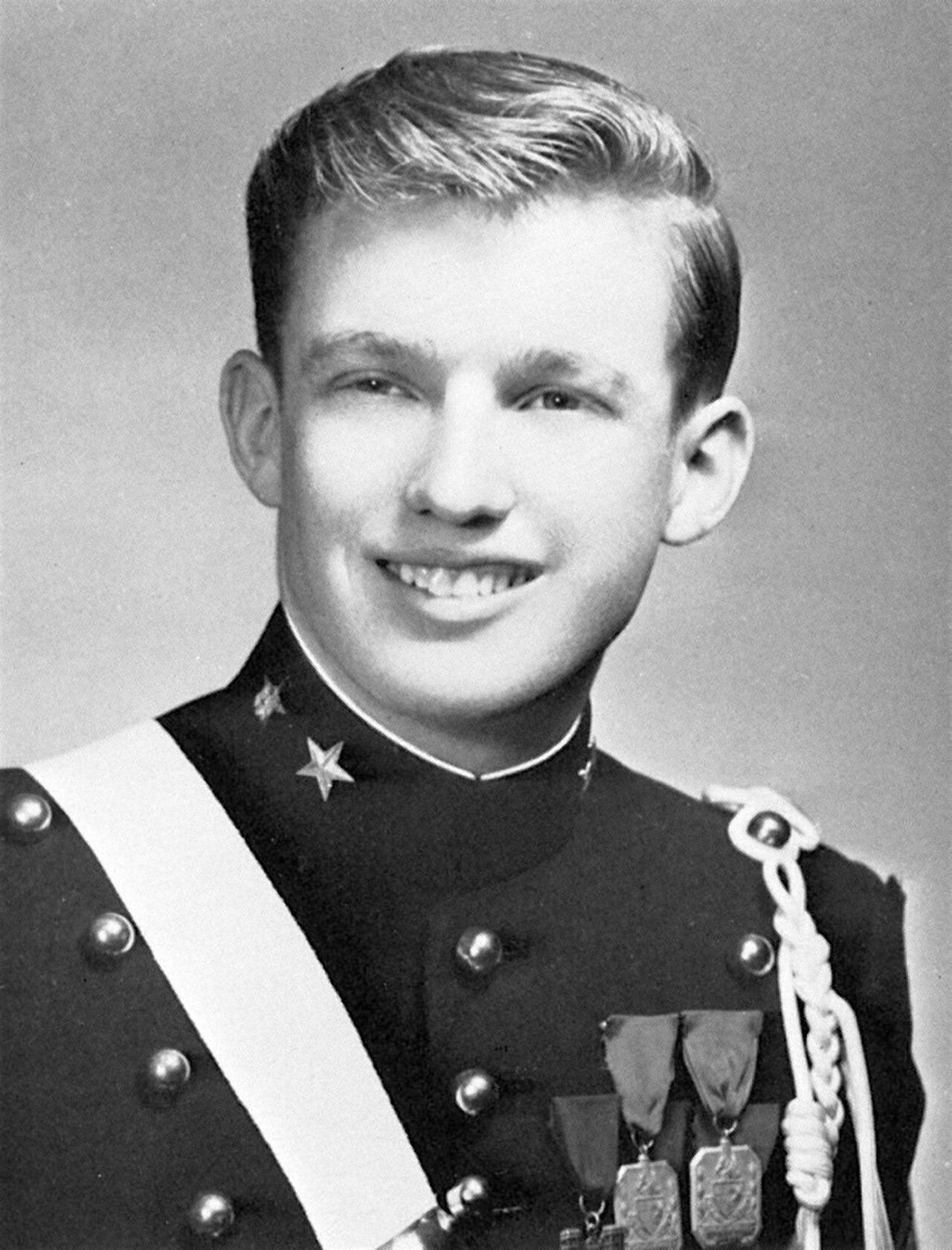 A black and white portrait of a young man in a military-style uniform with a white sash and medals.