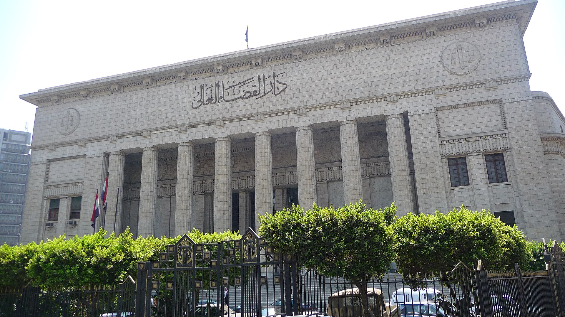A large, ornate stone building with columns and Arabic inscriptions above the entrance, surrounded by trees and a decorative fence.