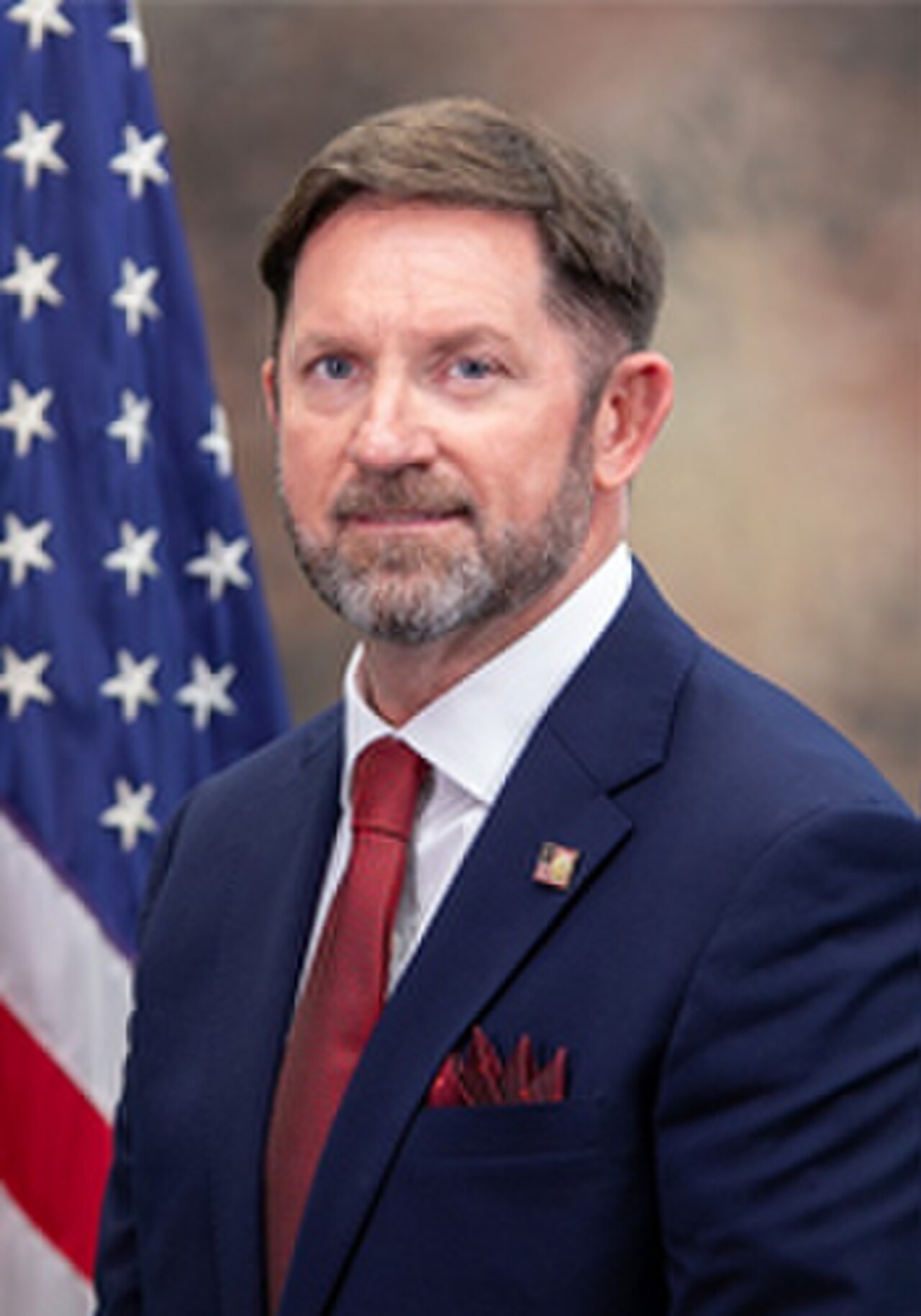 A man with short brown hair, a blue suit, red tie, and white shirt, with an American flag behind him.