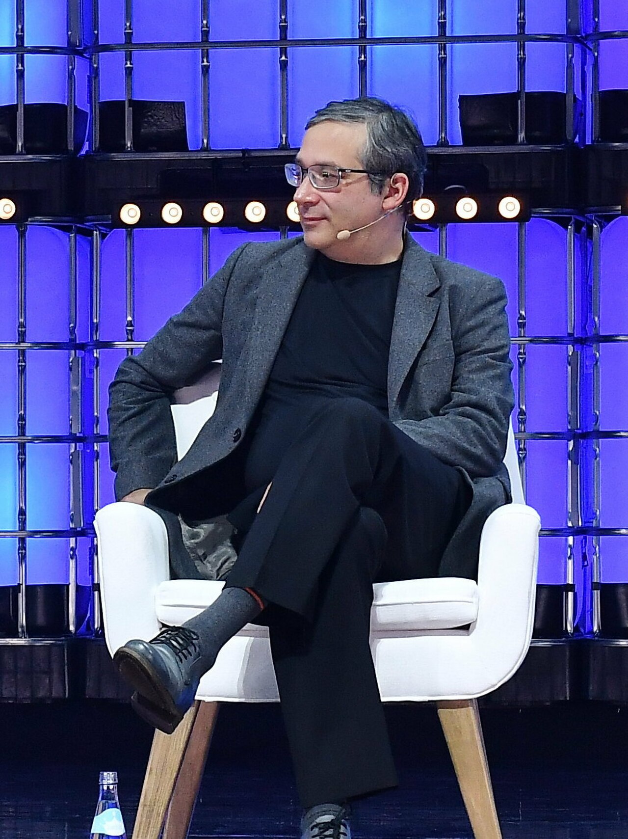 A man with short gray hair, wearing a gray blazer, black shirt, black pants, and black shoes, sits in a white chair with his right leg crossed over his left knee.