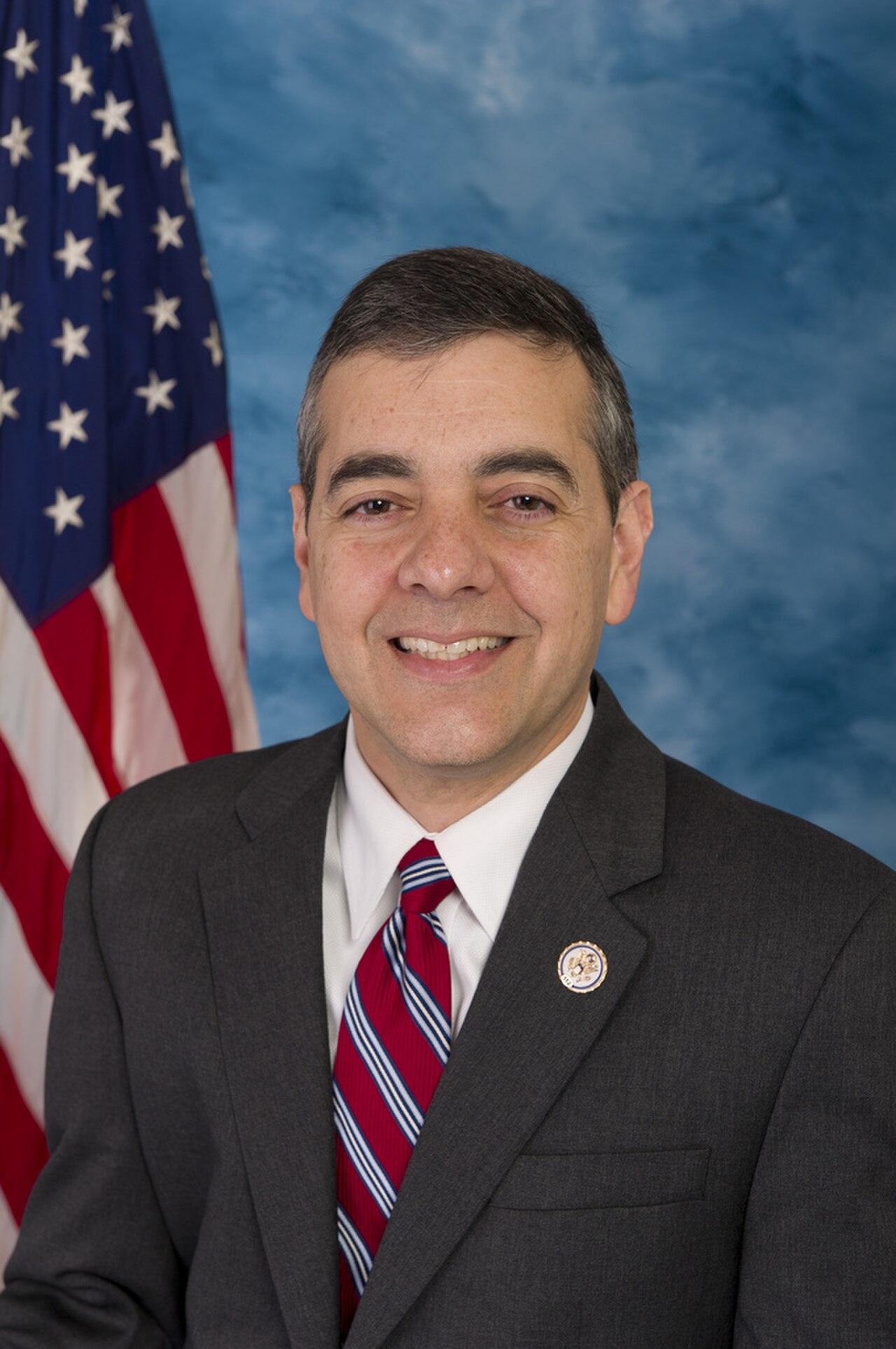 A head and shoulders portrait of a middle-aged man in a suit and tie, with an American flag behind him.