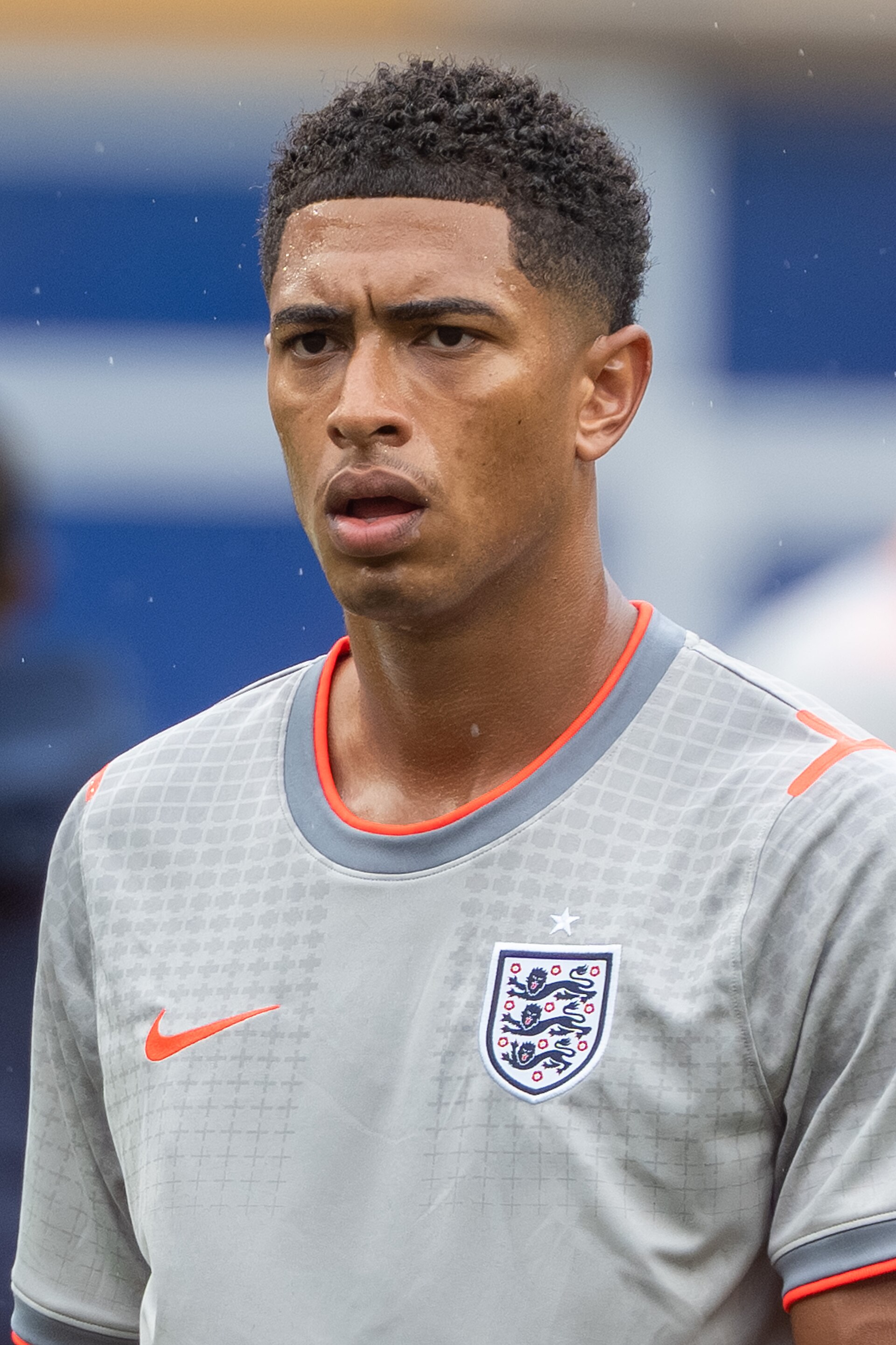 A young man with short dark hair wearing a gray soccer jersey with orange accents and the England national team crest.