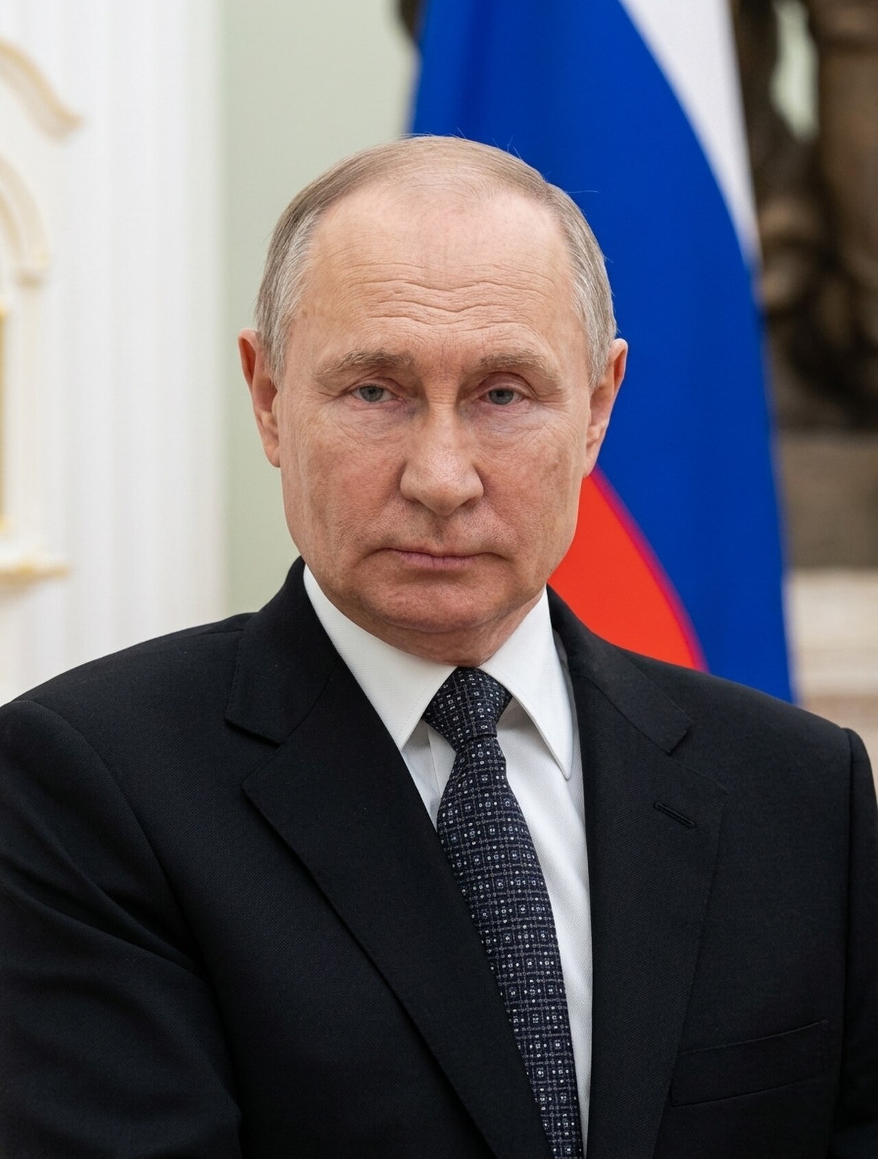 Vladimir Putin, a man with short gray hair, wearing a black suit, white shirt, and dark tie, stands in front of a blurred Russian flag.