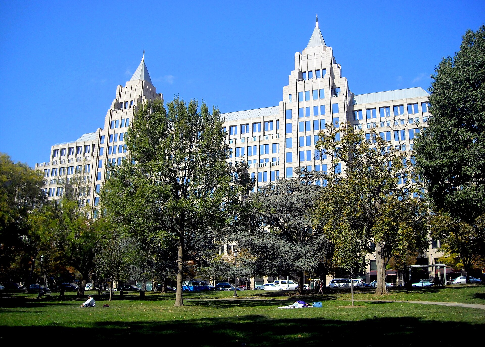 A large, light-colored building with two pointed towers and many windows stands behind a park with trees and people sitting on the grass.