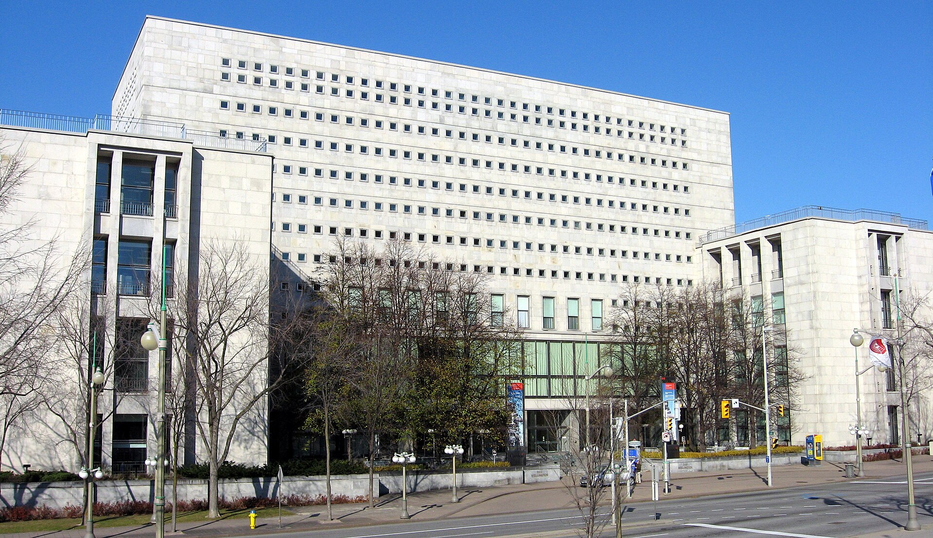 A large, multi-story white stone building with many windows, surrounded by trees and a street with traffic lights and signs.