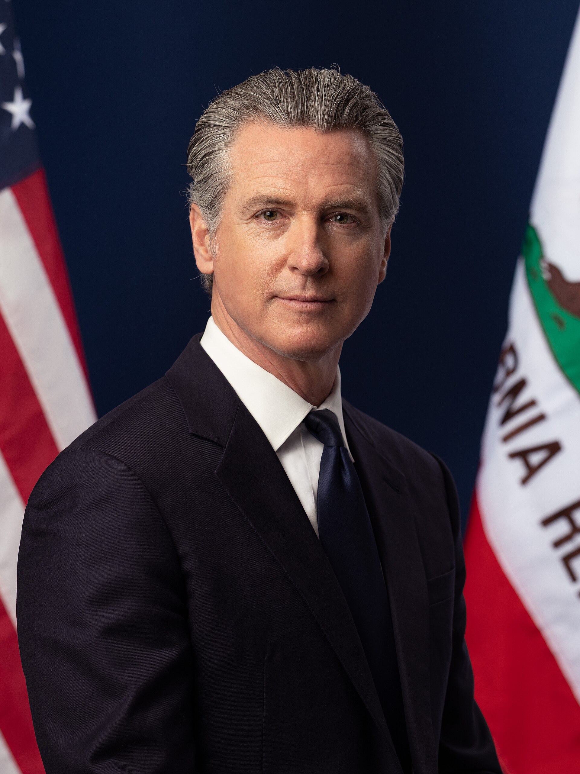 Gavin Newsom in a suit standing in front of two flags