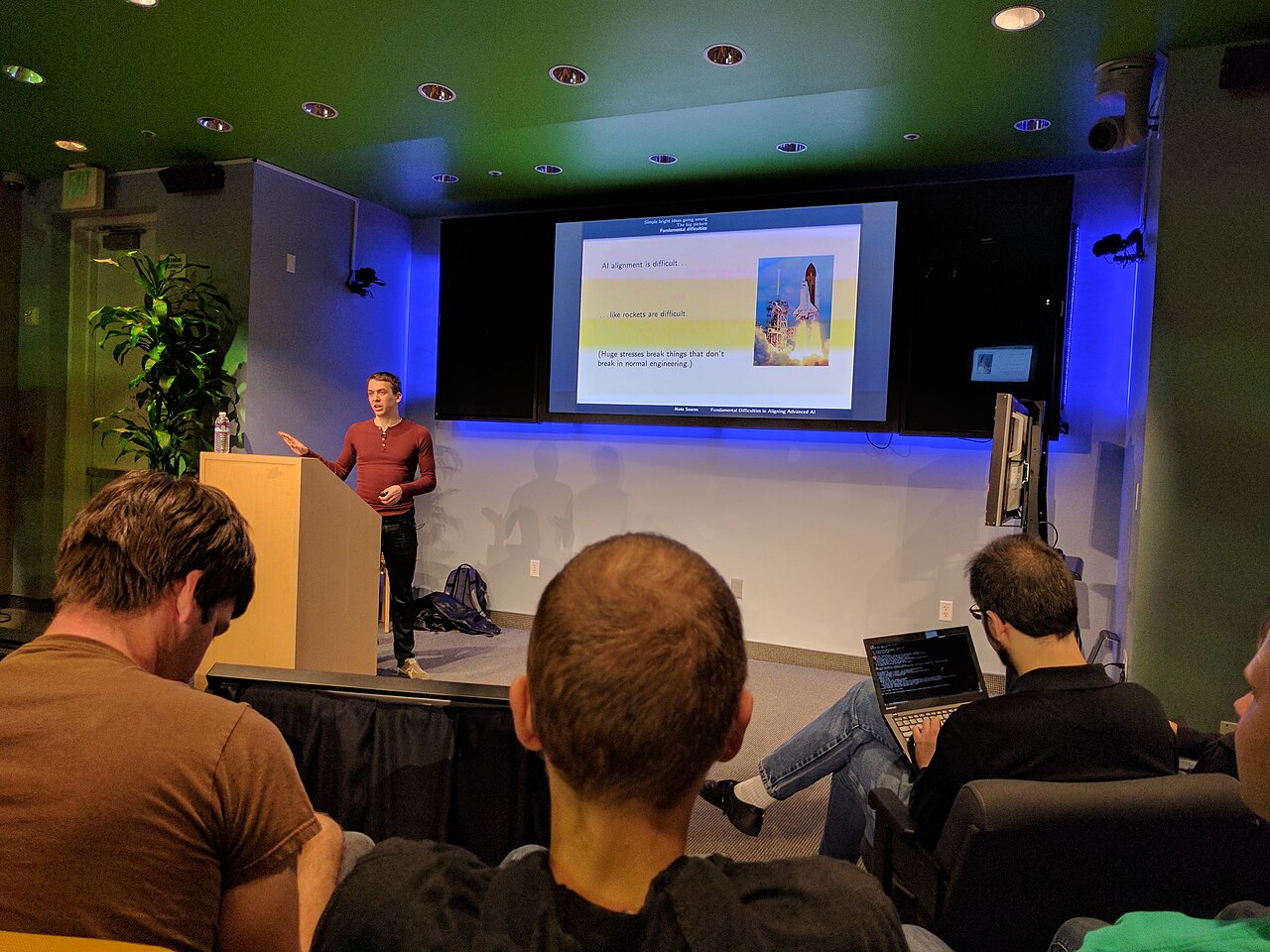 a man in a red shirt giving a presentation to a small audience in a conference room