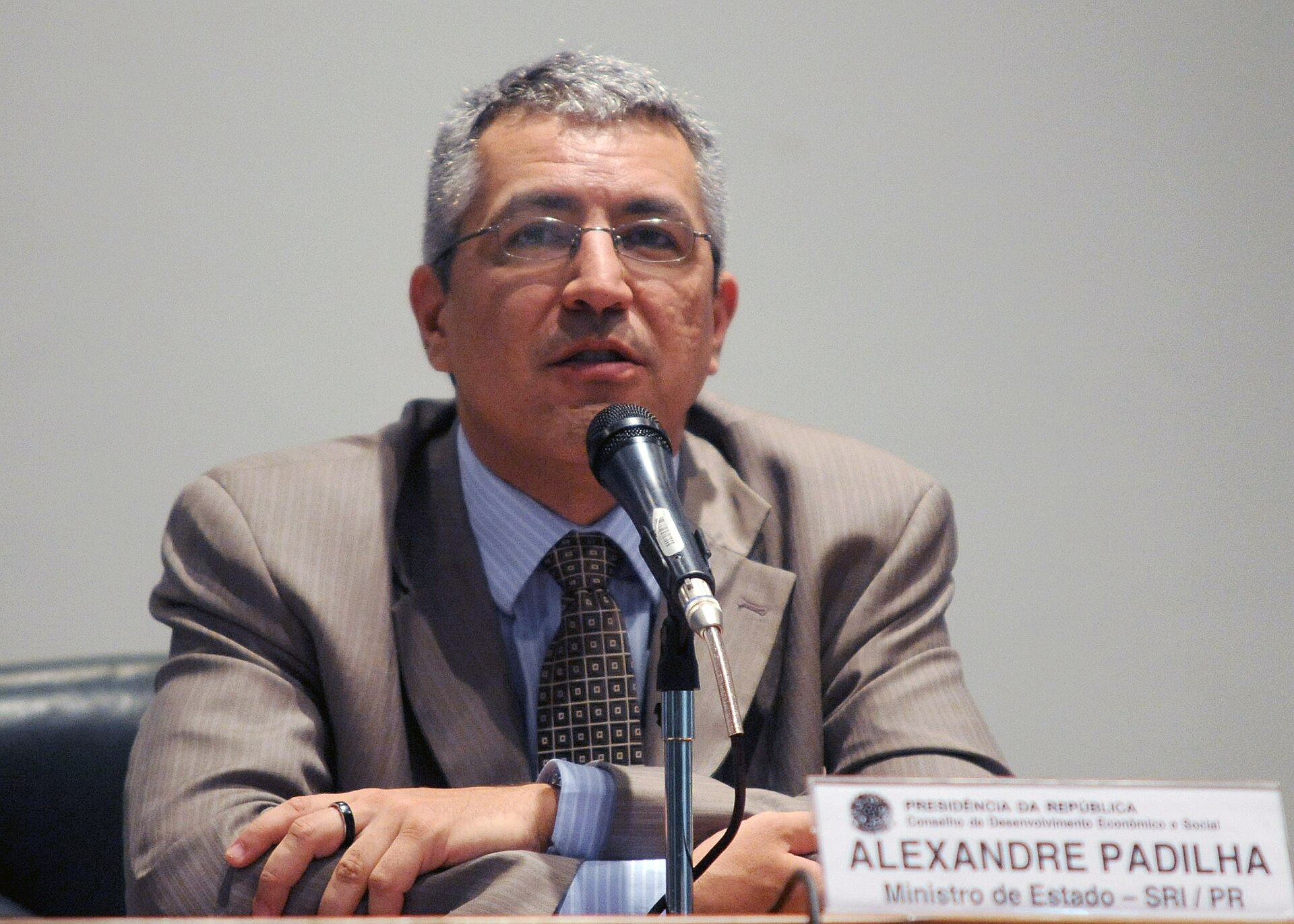 Alexandre Padilha, wearing a suit and tie, speaks into a microphone. A nameplate in front of him reads "PRESIDÊNCIA DA REPÚBLICA", "Conselho de Desenvolvimento Econômico e Social", "ALEXANDRE PADILHA", and "Ministro de Estado - SRI / PR".