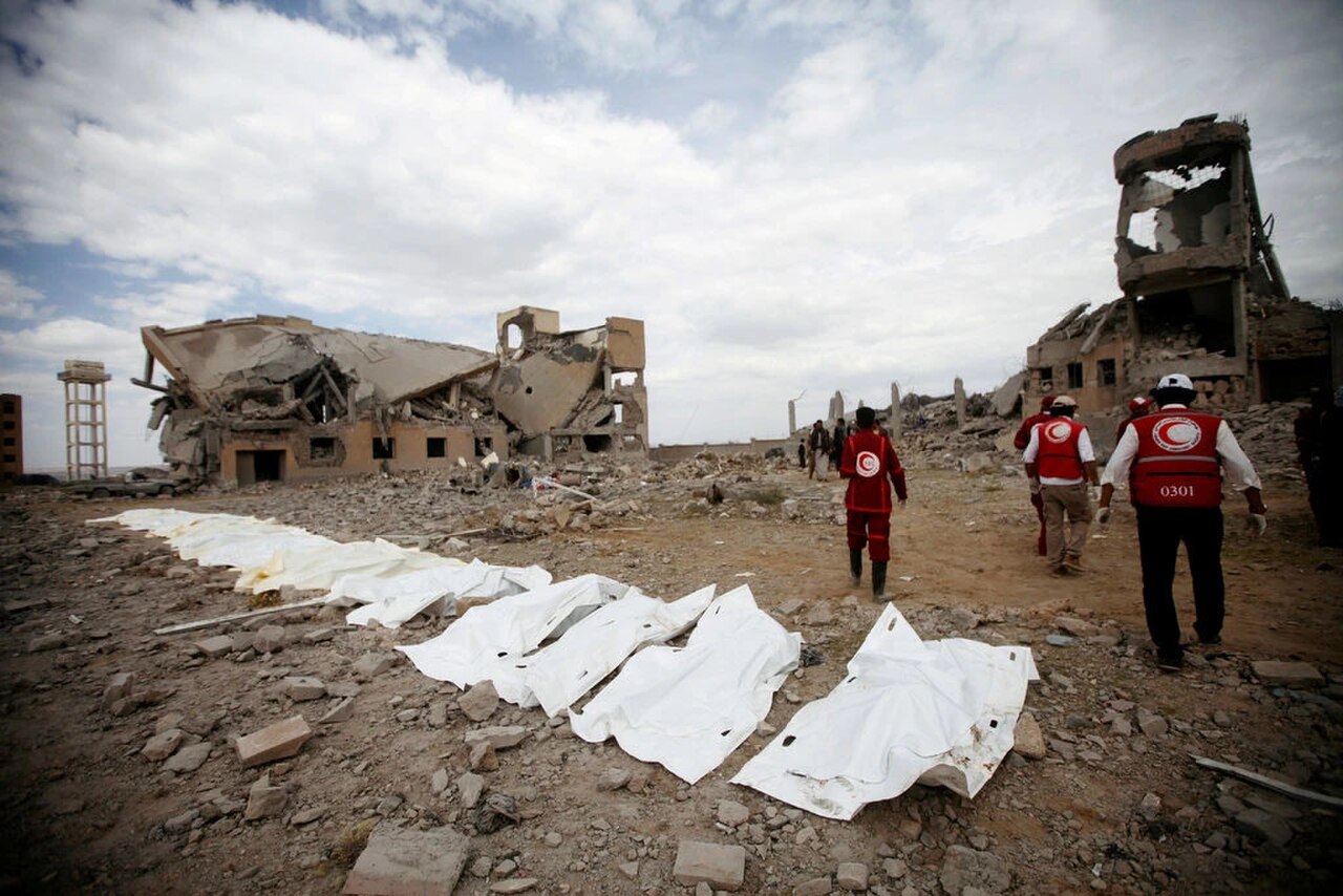 A scene of destruction with heavily damaged buildings and rubble. Several white body bags are laid out on the ground in the foreground. In the middle ground, several people wearing red vests with a white crescent emblem are walking away from the camera, towards the damaged structures.