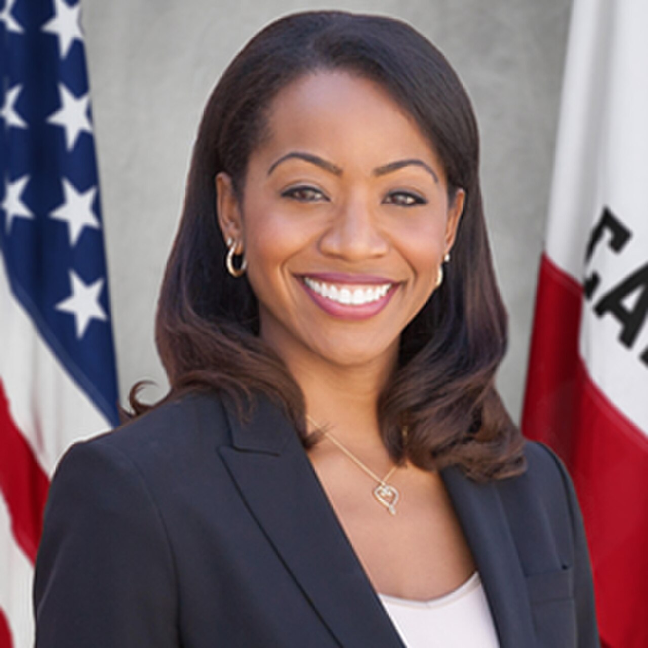 A smiling woman with dark hair and a black blazer standing in front of an American flag and a California state flag.