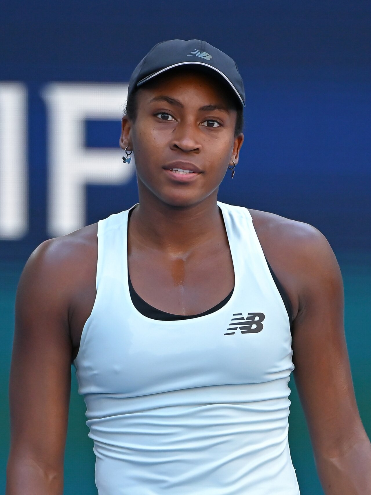 Coco Gauff, a young black woman, wearing a white New Balance tank top and a black cap with a New Balance logo, standing in front of a blue and green background with a partial white letter.