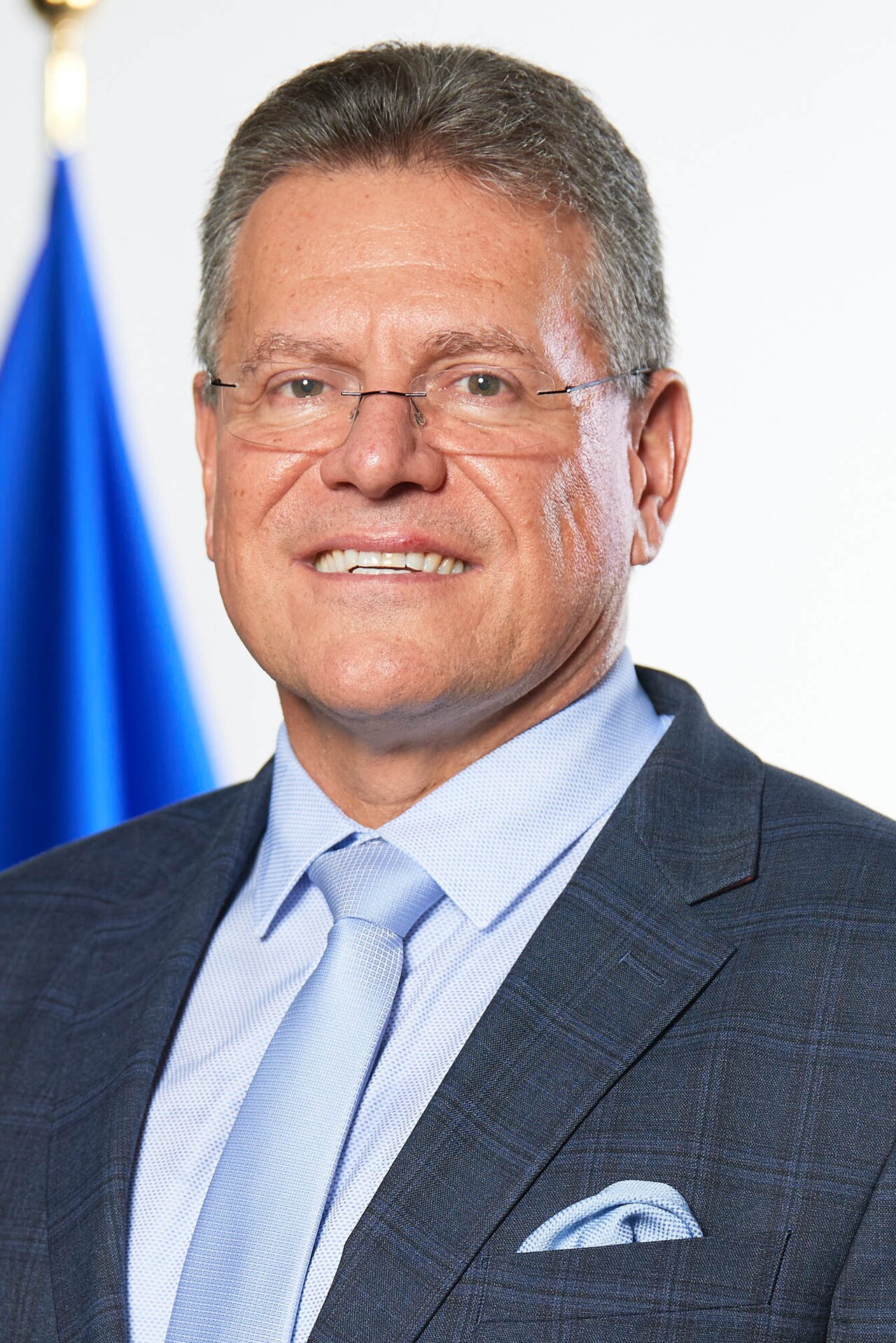 A man with short gray hair wearing a blue suit, light blue shirt, and light blue tie, with a blue flag in the background.