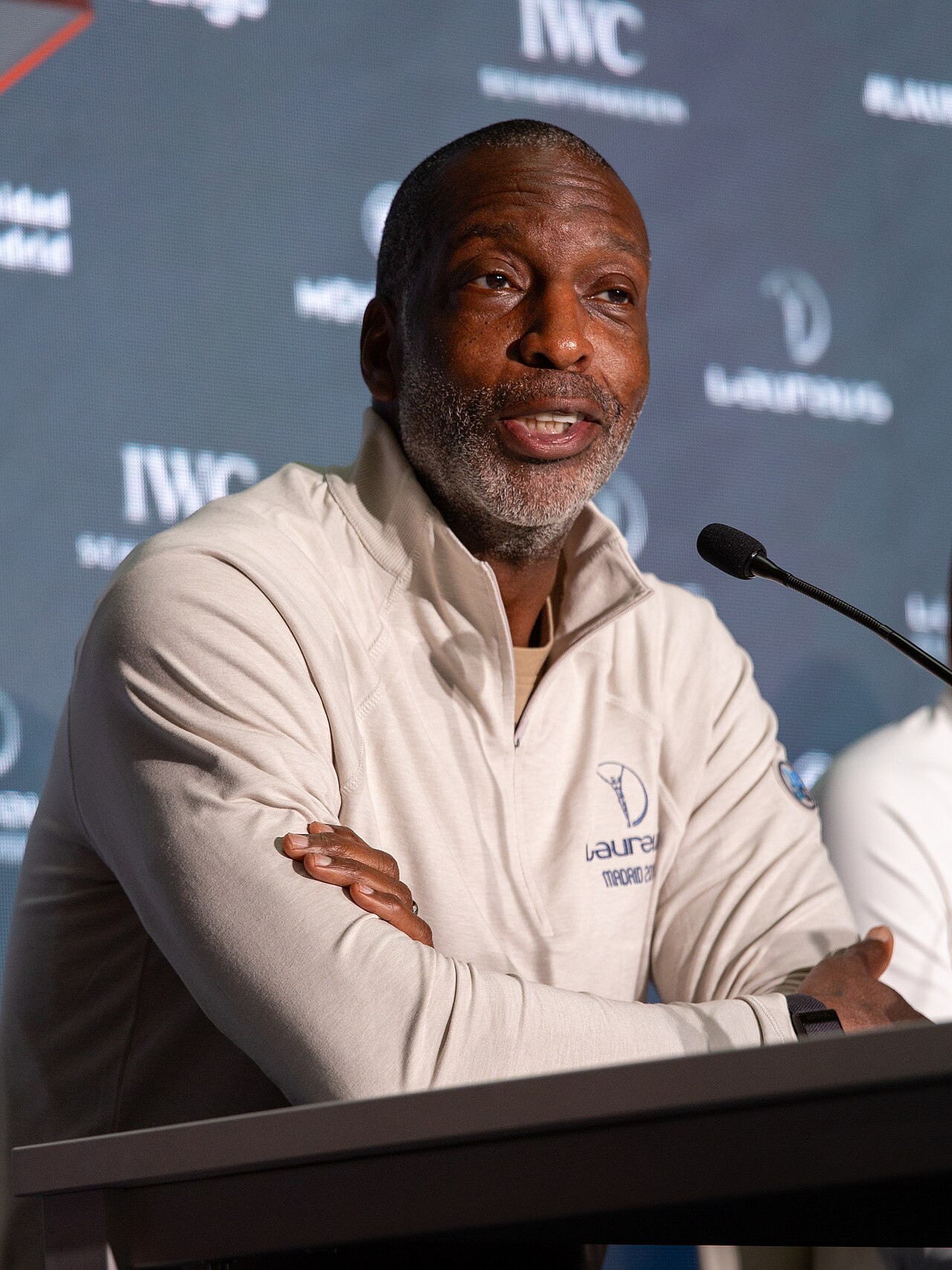 Michael Johnson, a dark-skinned man with short black hair and a graying beard, wearing a beige quarter-zip pullover with a logo on the left chest, sitting at a podium with his arms crossed.