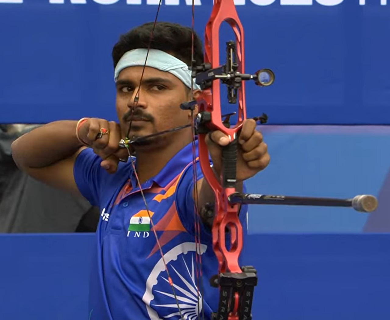 A man in a blue jersey with an orange and white design and the Indian flag on it holds a red bow and aims an arrow.