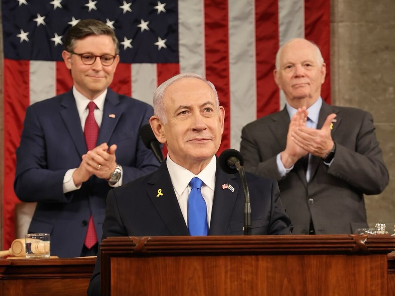 A man in a dark suit, blue tie, and yellow ribbon speaks at a podium, with two men in suits applauding behind him. An American flag hangs in the background.