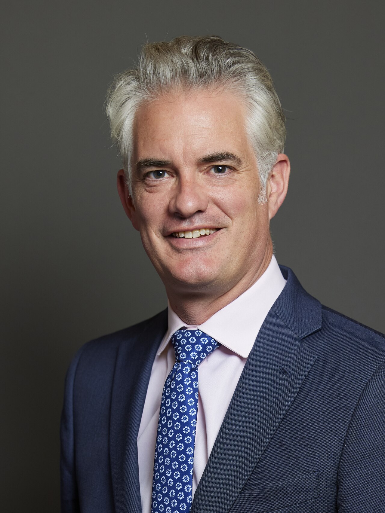 A middle-aged man with short gray hair, wearing a blue suit, white shirt, and blue tie with white flowers, against a gray background.