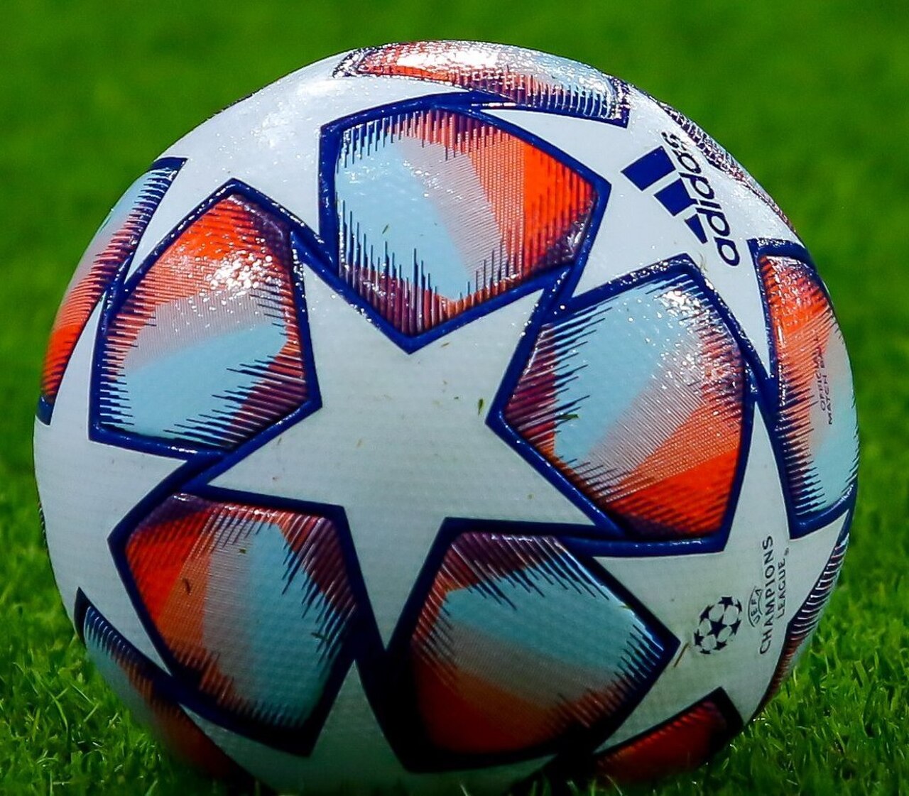 A close-up shot of a white Adidas Champions League soccer ball with orange and blue star-shaped panels, resting on green grass.