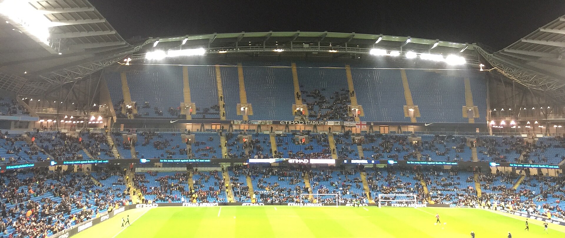 A wide shot of the Etihad Stadium at night, with blue seats and yellow accents visible. The stadium is illuminated by floodlights, and a crowd is seated in the stands. The words "ETIHAD STADIUM" are visible on the upper tier.