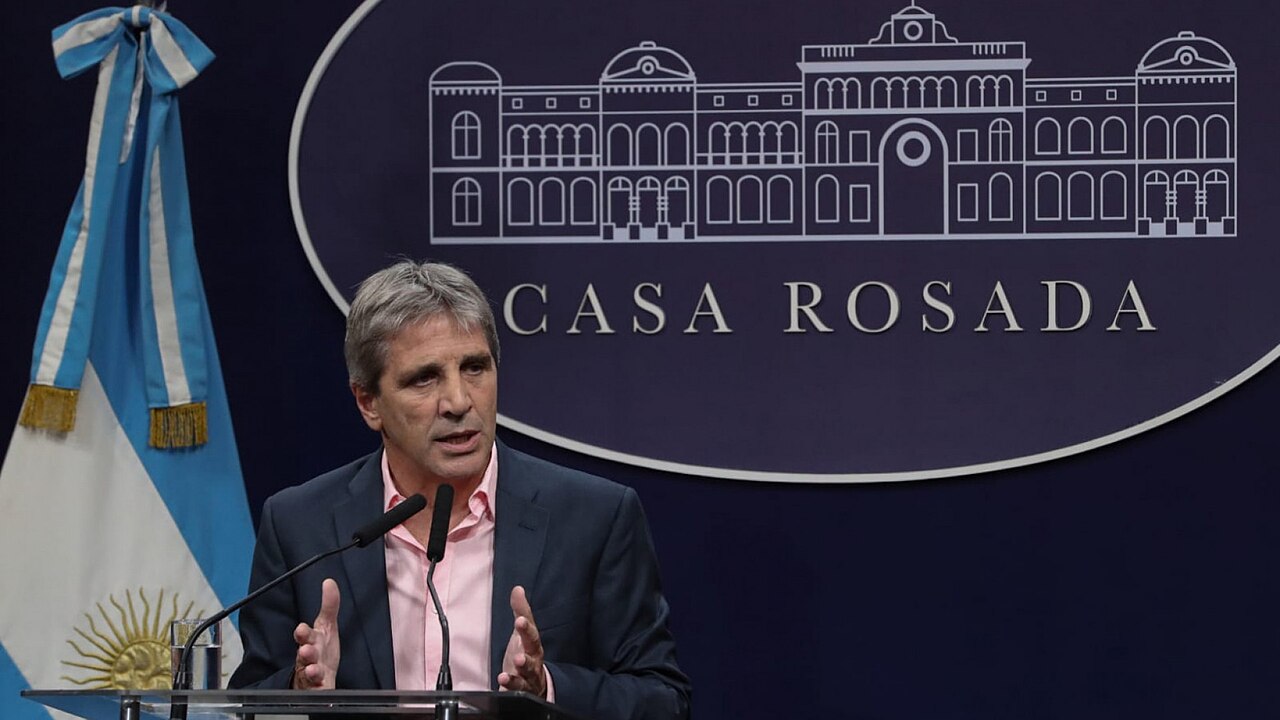 A middle-aged man with graying hair in a suit jacket and pink shirt speaking at a podium with two microphones, in front of a dark blue backdrop with a Casa Rosada logo and an Argentine flag.