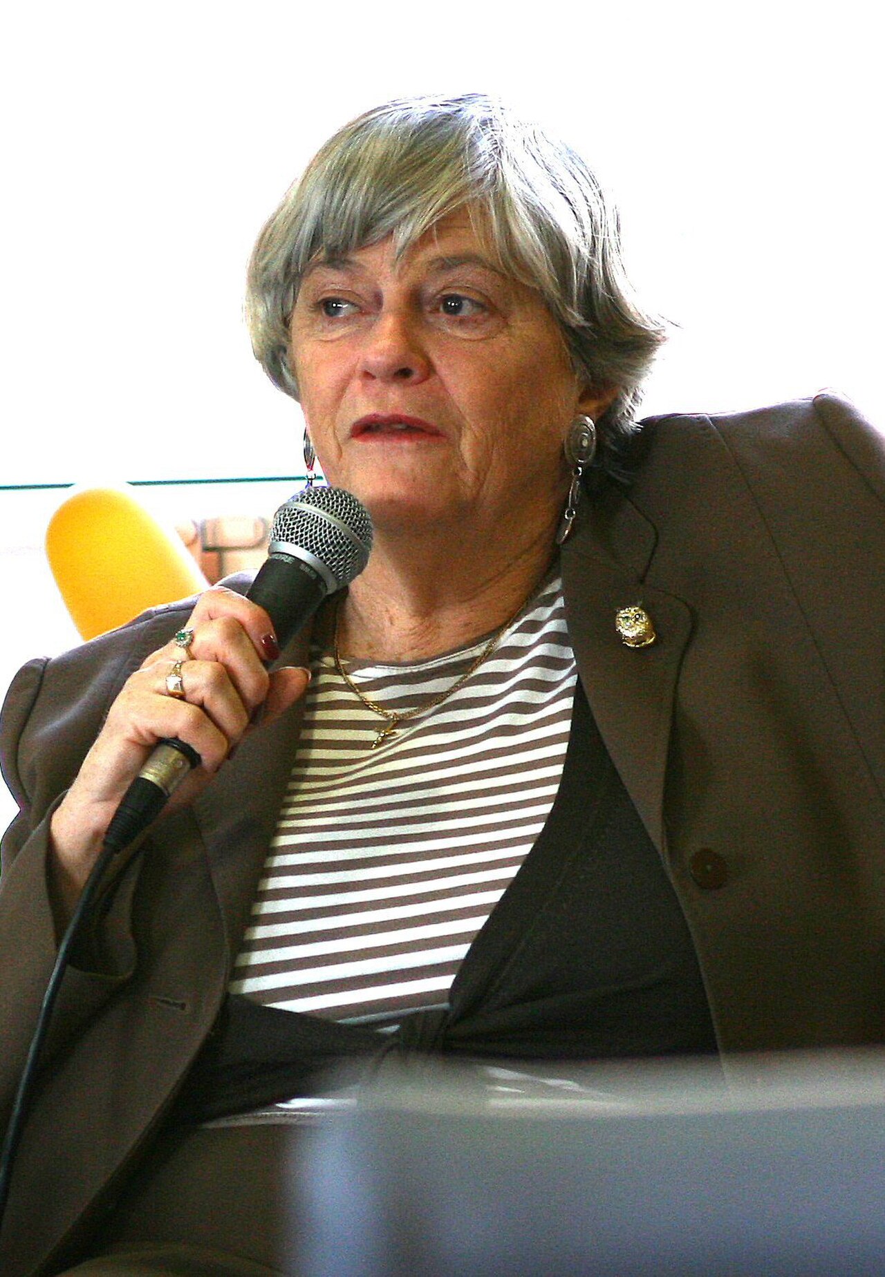 A woman with short gray hair, wearing a brown blazer, striped shirt, and jewelry, holding a microphone.