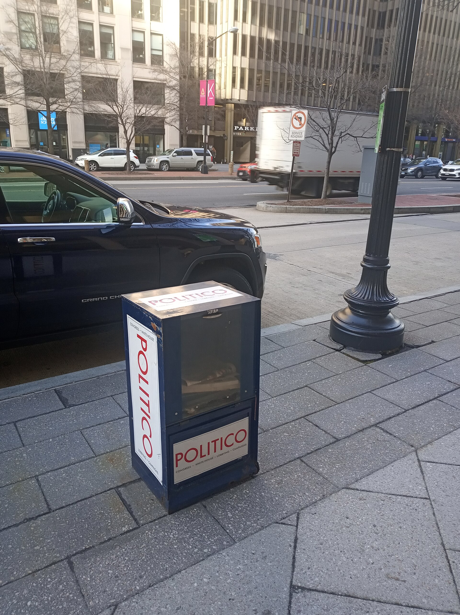 A blue Politico newspaper box stands on a sidewalk next to a dark blue car. The box has the Politico logo on its sides and front.