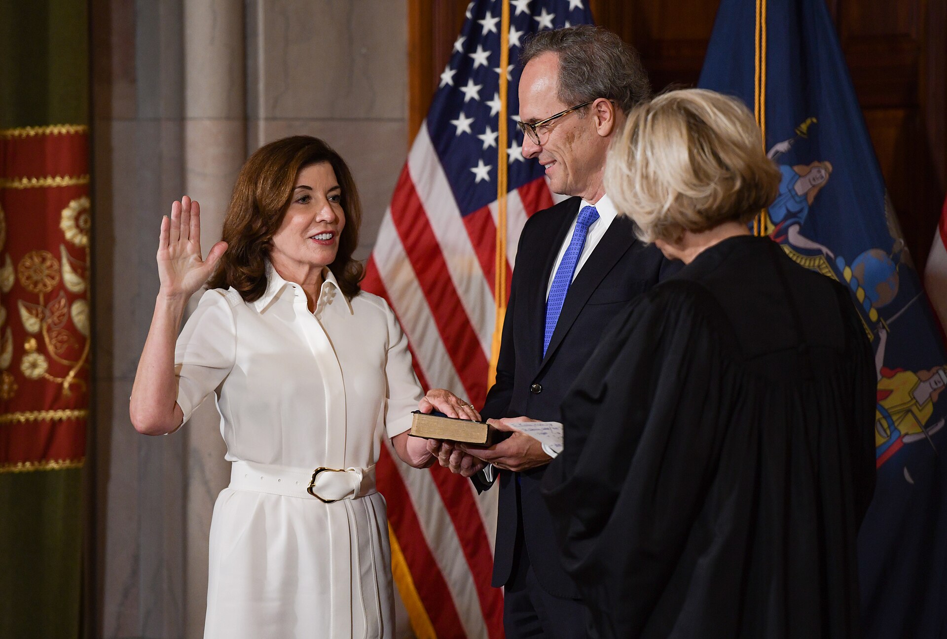A woman in a white dress raises her right hand while placing her left hand on a bible held by a man in a suit. Another person in a black robe stands to the right.