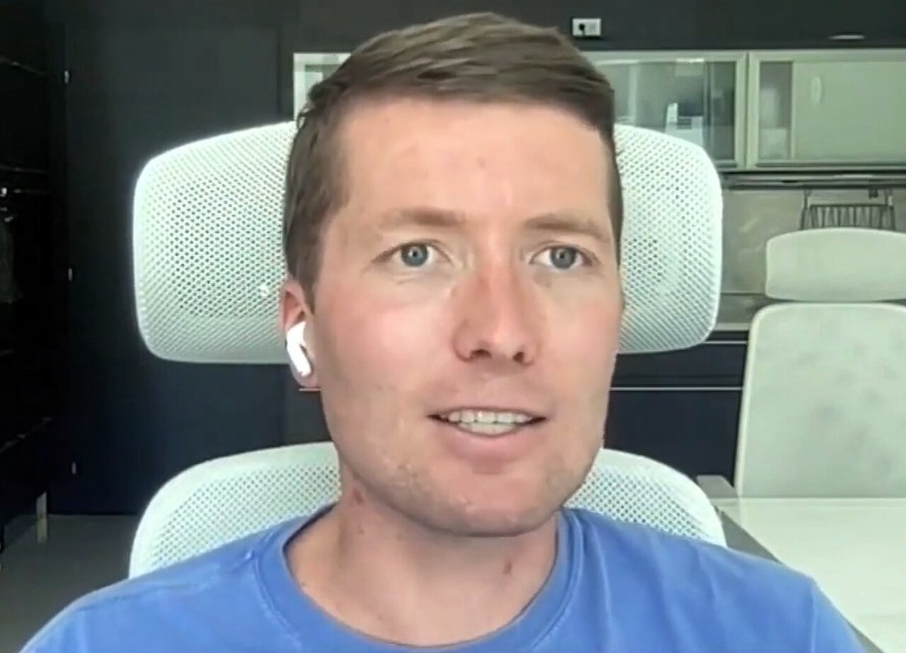 A man with short brown hair, blue eyes, and a blue shirt, wearing white earbuds, sitting in a white office chair.